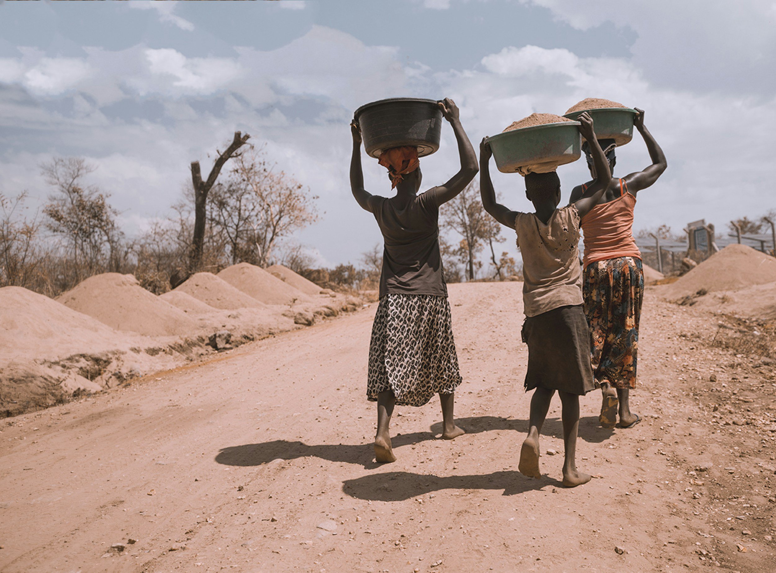 Three women in Uganda walk on a dirt road carrying large baskets on their heads, in a dry landscape with bare trees and mounds of dirt.