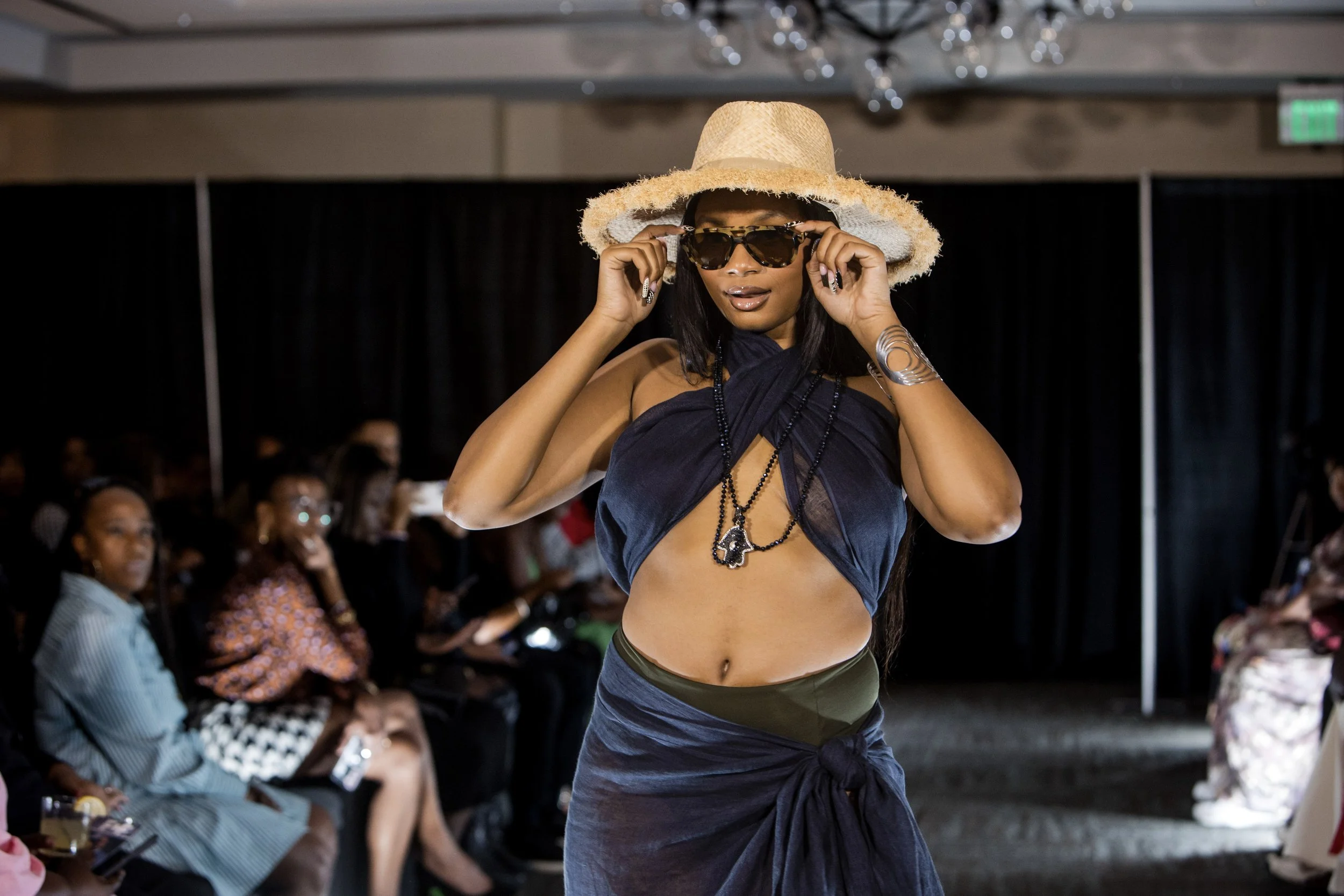 Woman on runway wearing a large straw hat, sunglasses, a dark wrap top, and a skirt with a tied knot, during a fashion show.