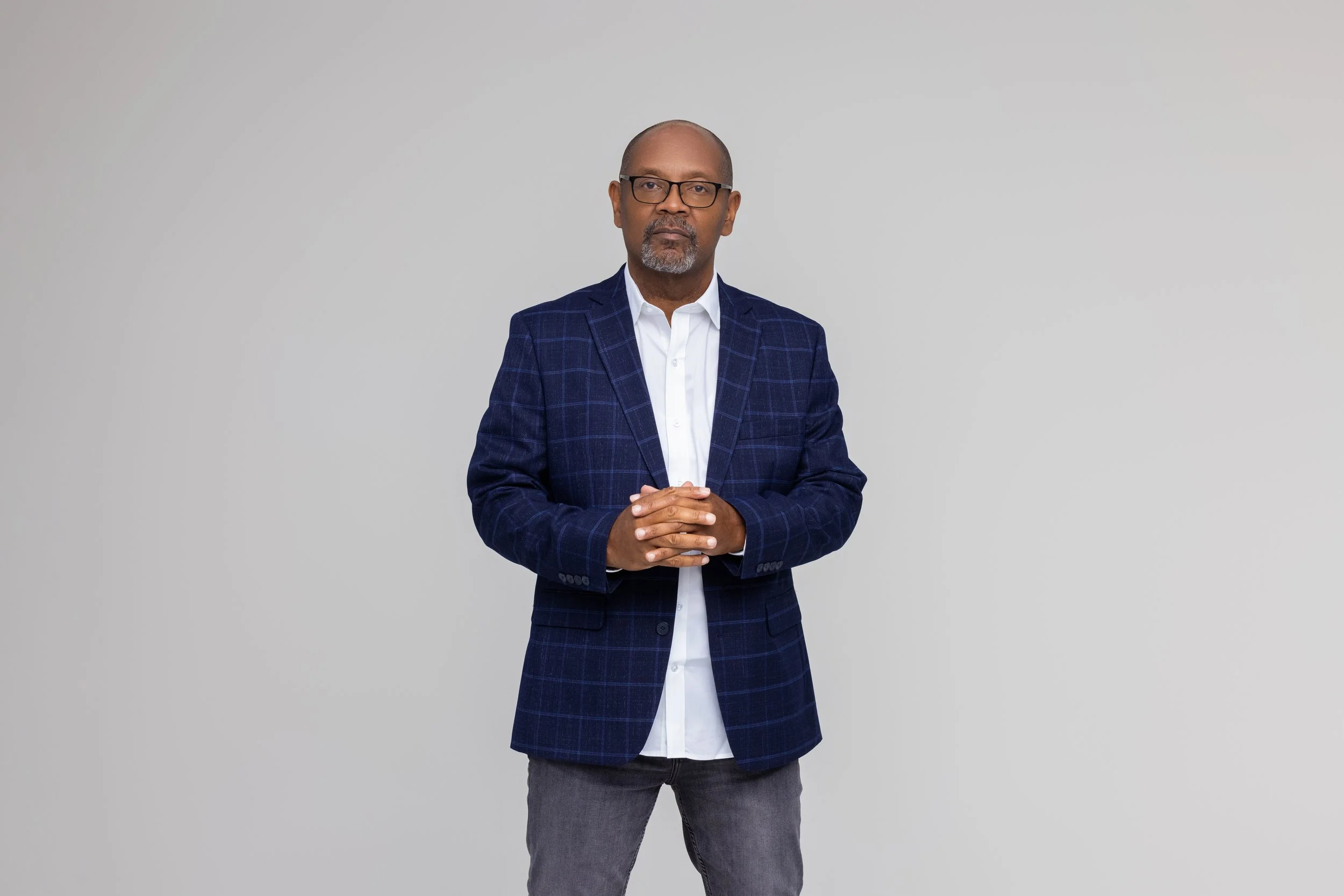 A middle-aged man with glasses, a beard, and a bald head wearing a dark blue checkered blazer, white shirt, and gray jeans, standing with hands clasped in front of a plain gray background.