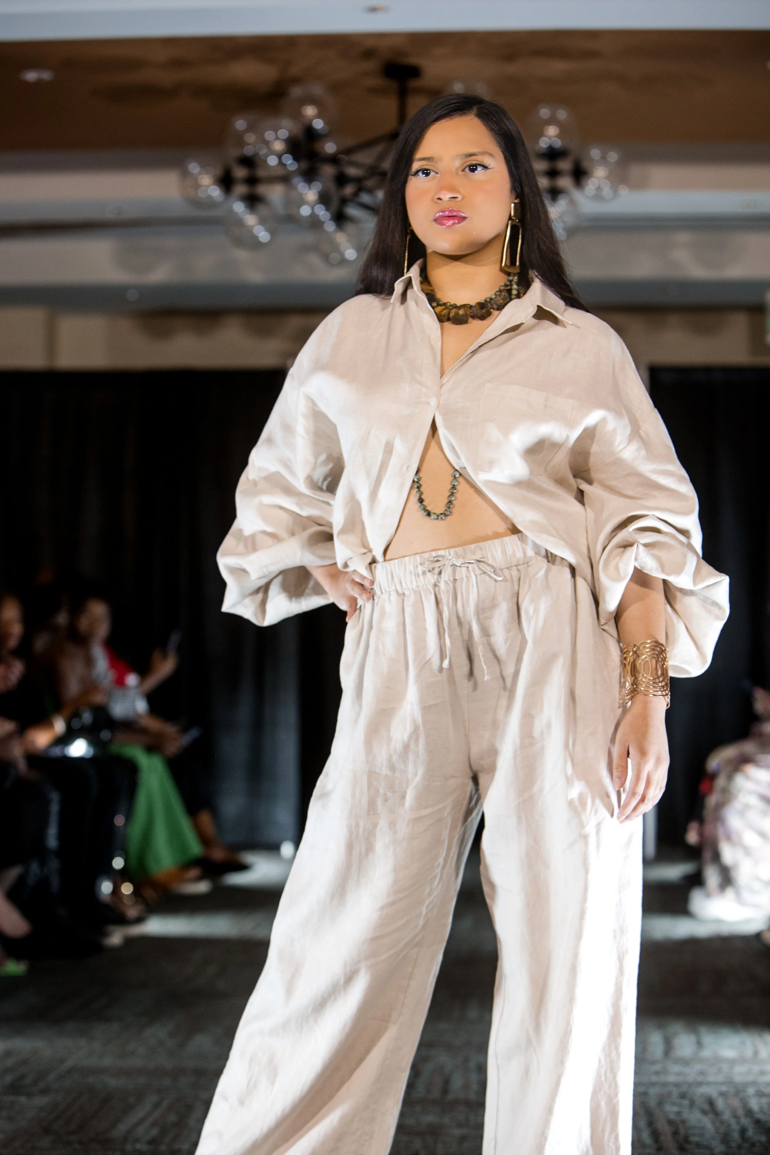 A woman walking on a runway during a fashion show, wearing beige wide-leg pants and a matching loose shirt, accessorized with large gold earrings, a beaded necklace, and a gold bracelet.