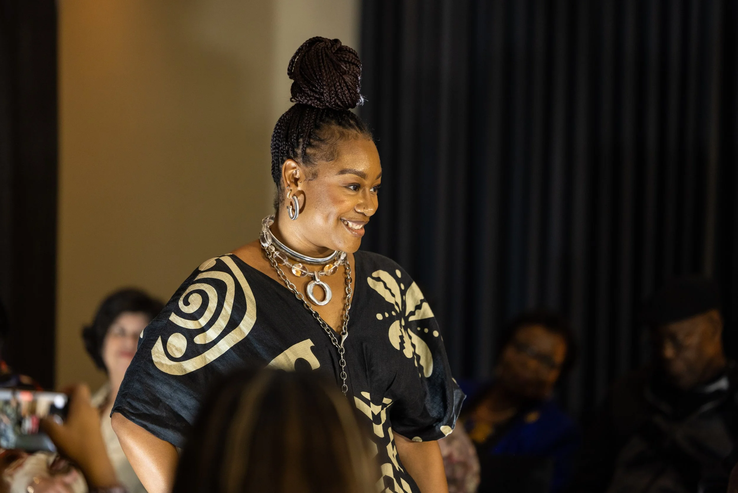A woman with braided hair styled in a high bun, wearing large earrings and layered silver necklaces, smiling during a social event or gathering.