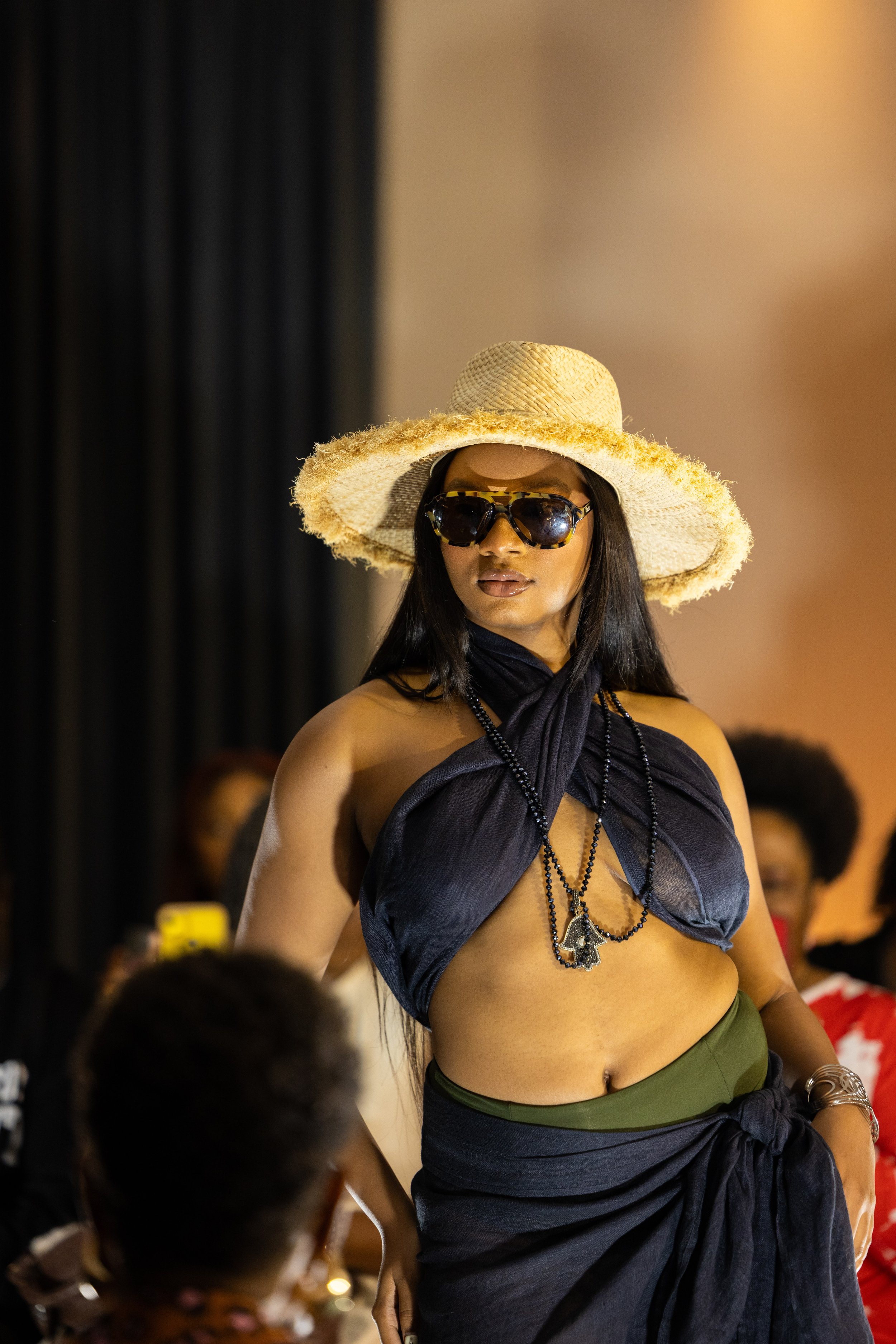 Woman wearing a wide-brim straw hat, dark sunglasses, a black scarf top, and jewelry, standing in front of a crowd indoors.