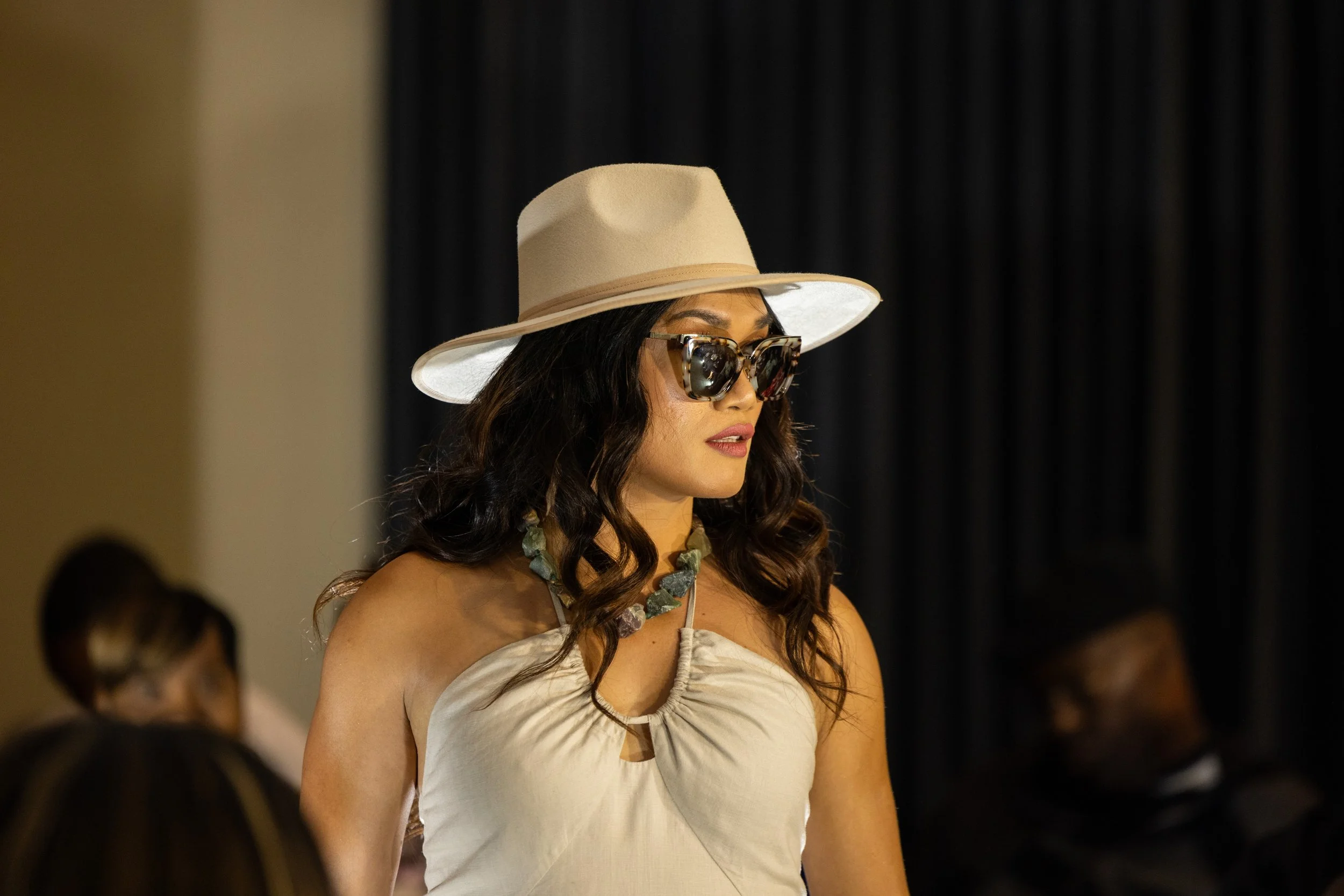 A woman wearing a beige hat, large sunglasses, a beige top, and a beaded necklace, standing in a dimly lit room with dark curtains in the background.
