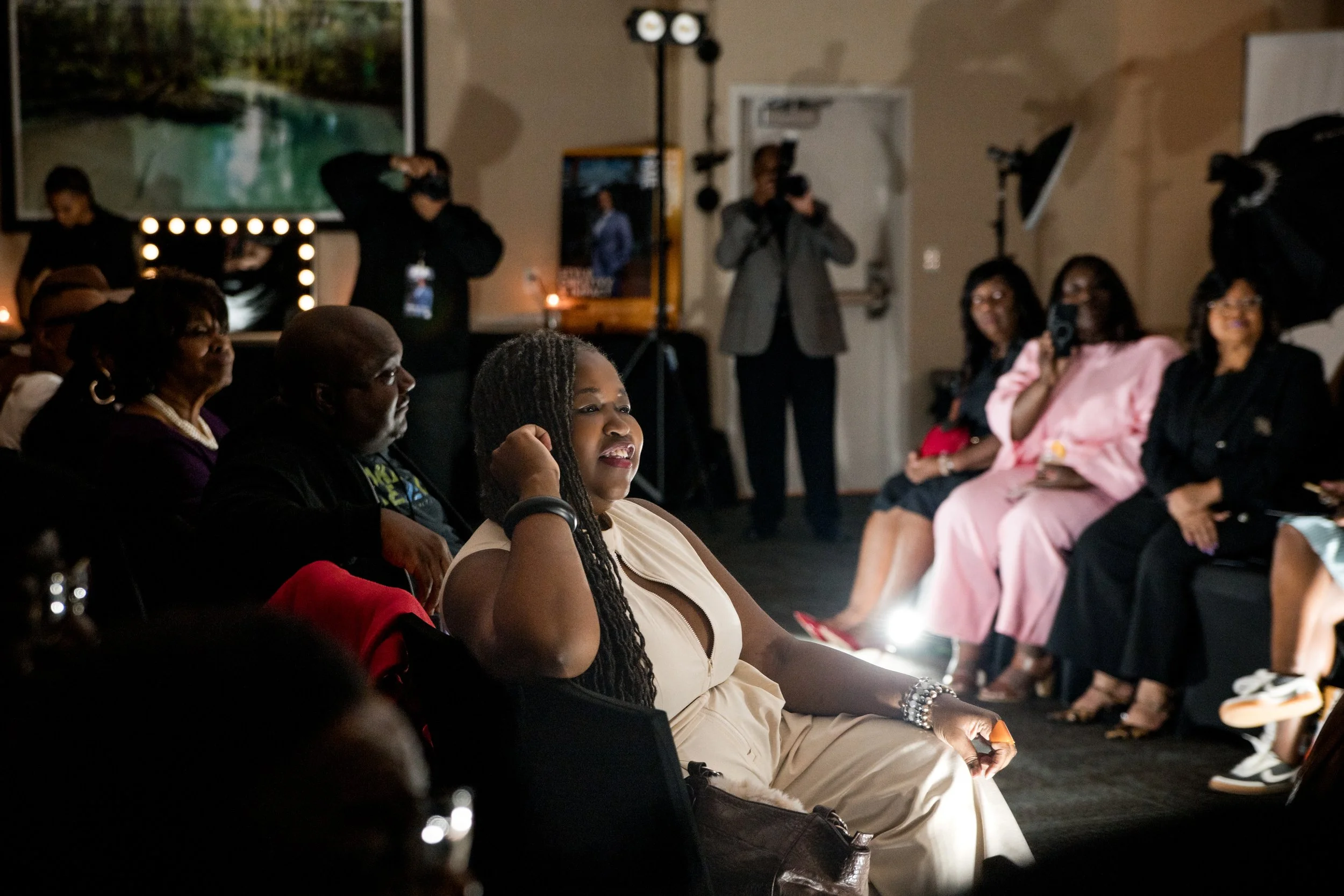 Women sitting in a dimly lit room, listening to a speaker or presentation, with cameras and photographers capturing the event.