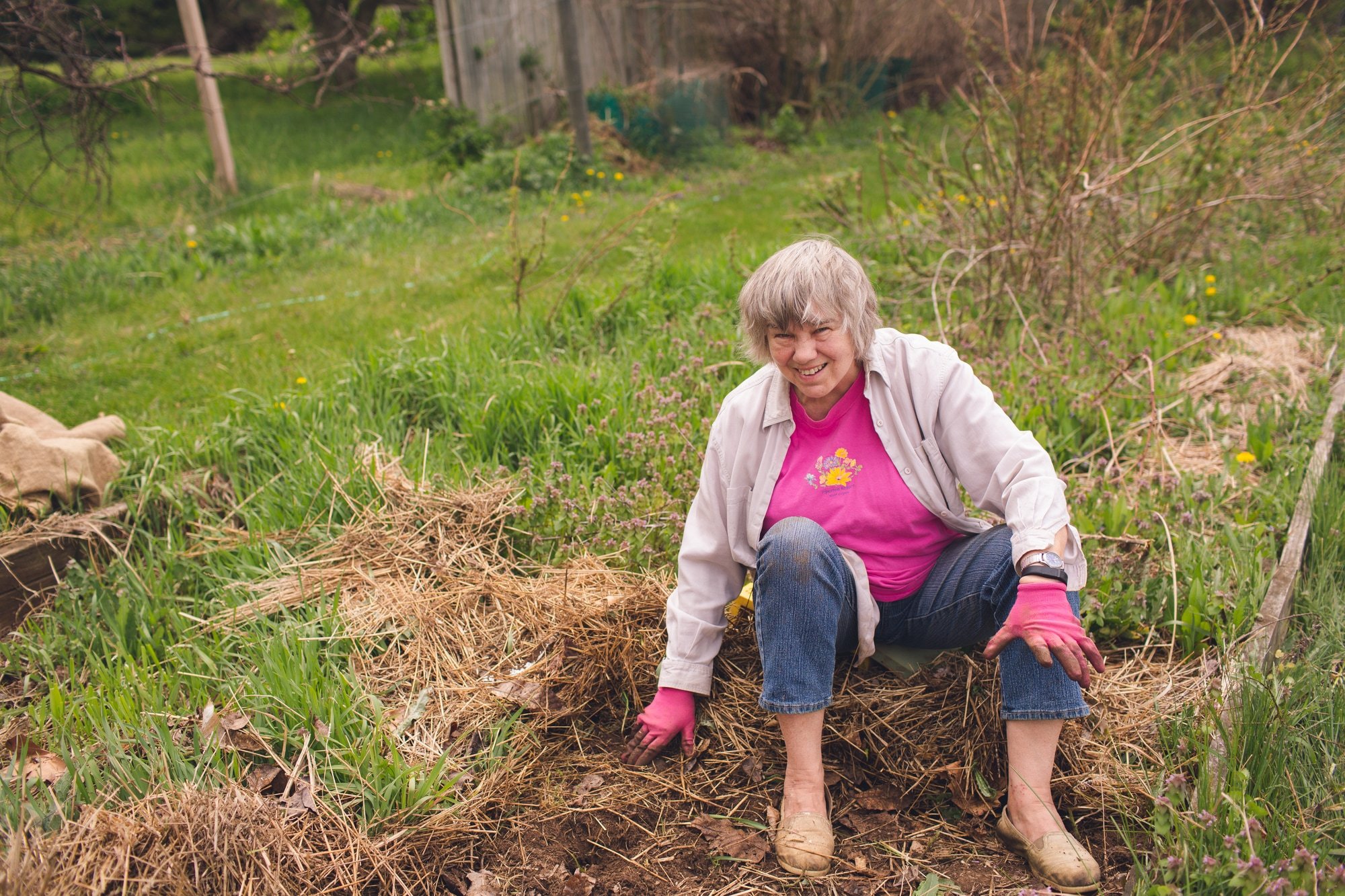 A woman sitting on a pile of straw smiles at the camera while doing some garden work.