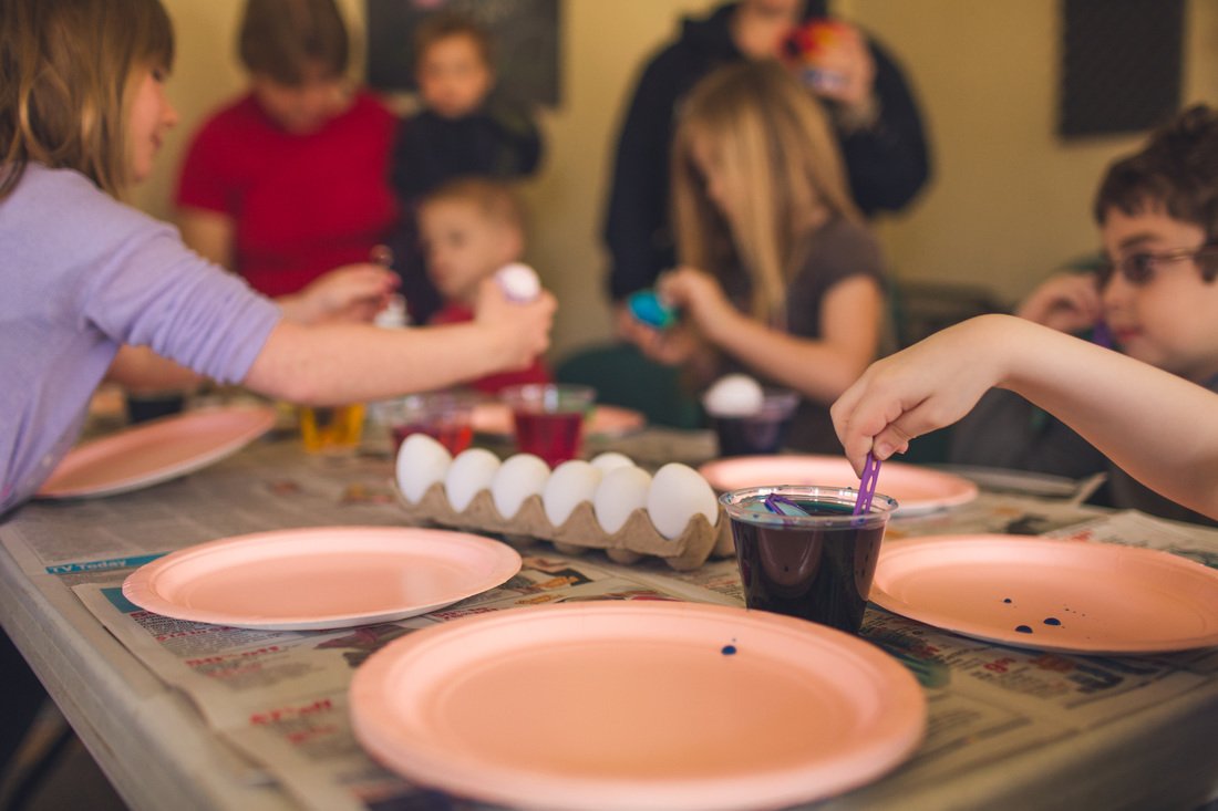 Children at a table protected by newspapers with eggs, cups of dye, and paper plates.