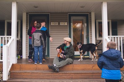 A small group of children and adults with a dog socializing on a front porch. A man sits on the top step playing a guitar.