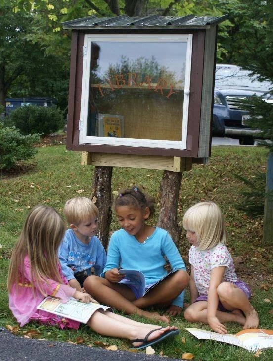 Children sitting in the grass reading together under a little library.
