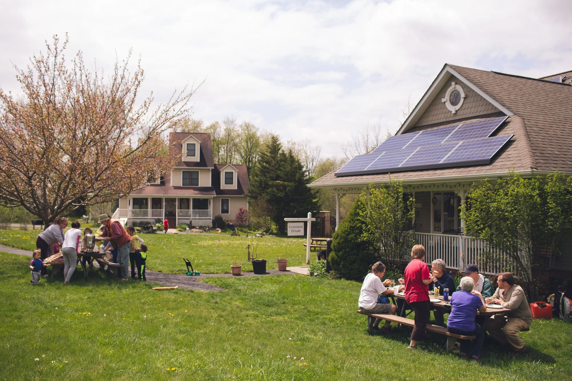 Children and adults sitting at a picnic table in front of a community building. The building has solar panels installed and a "Common House" sign.