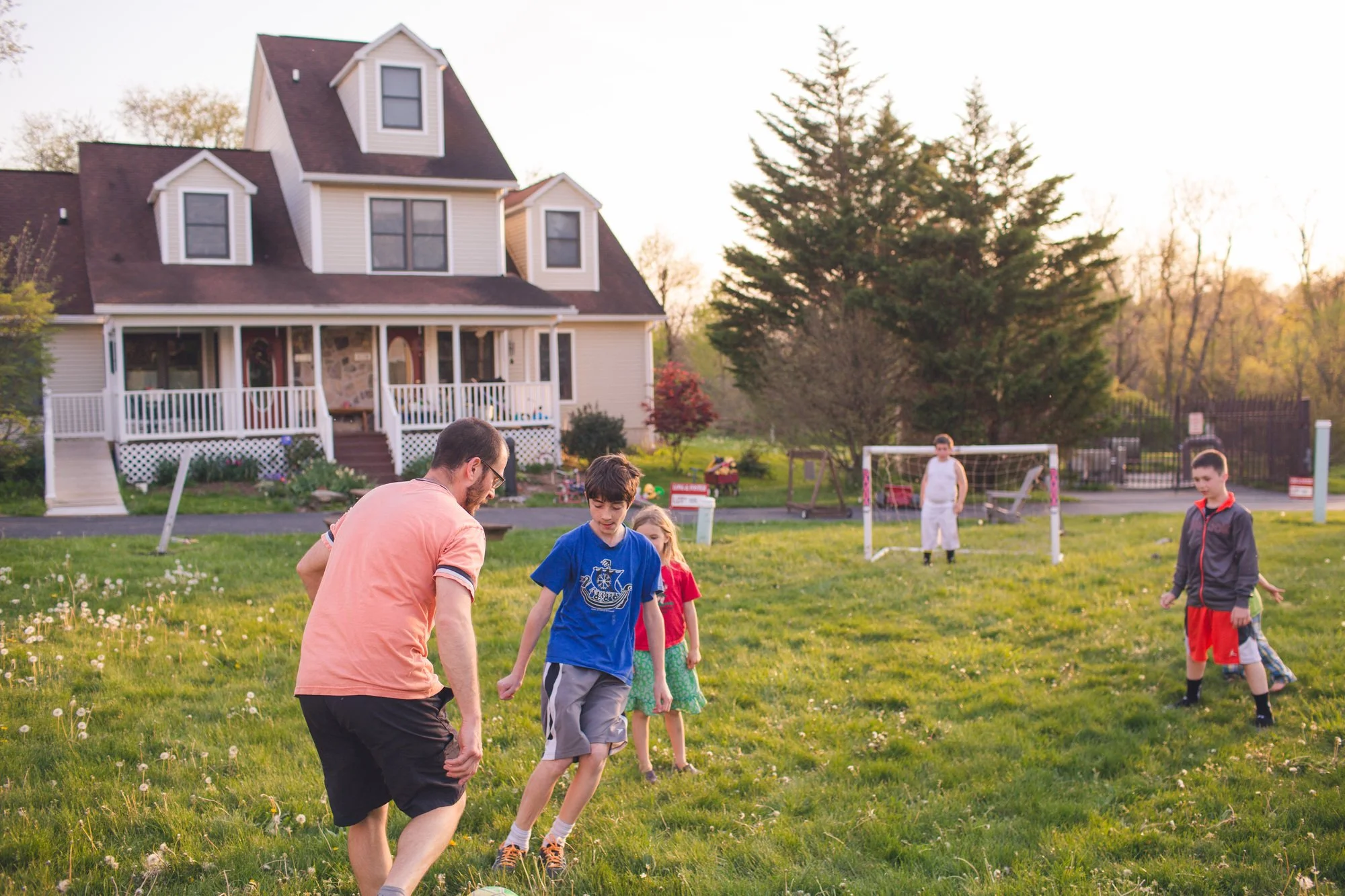 An adult and group of children playing soccer in the grass field in front of a home.