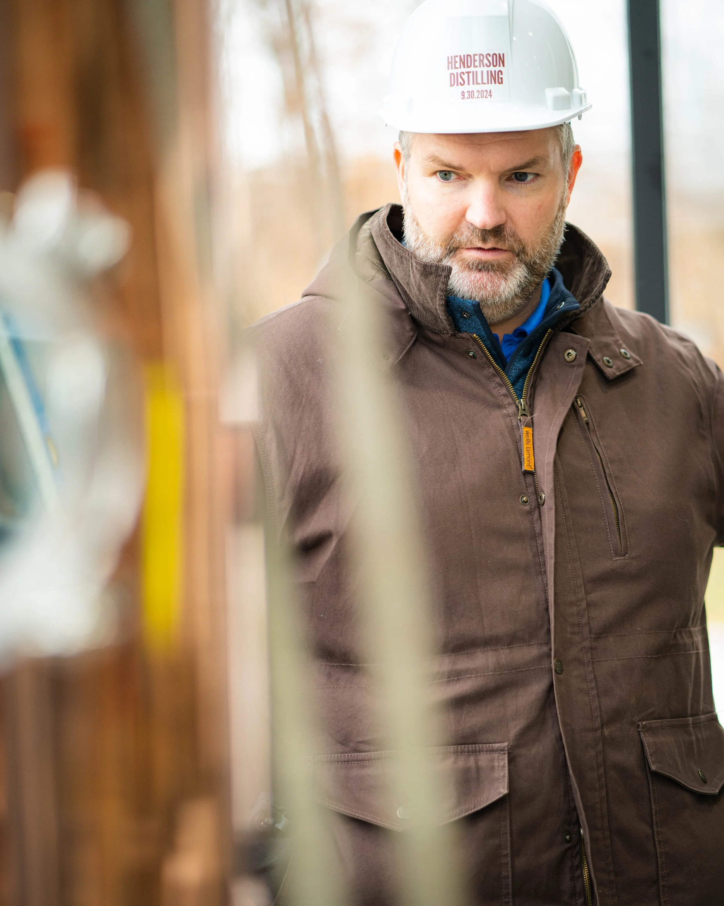 A man wearing a hard hat labeled 'HENDERSON DISTILLING 9.30.2024' and a brown jacket looking to the side inside a building with a blurred background.