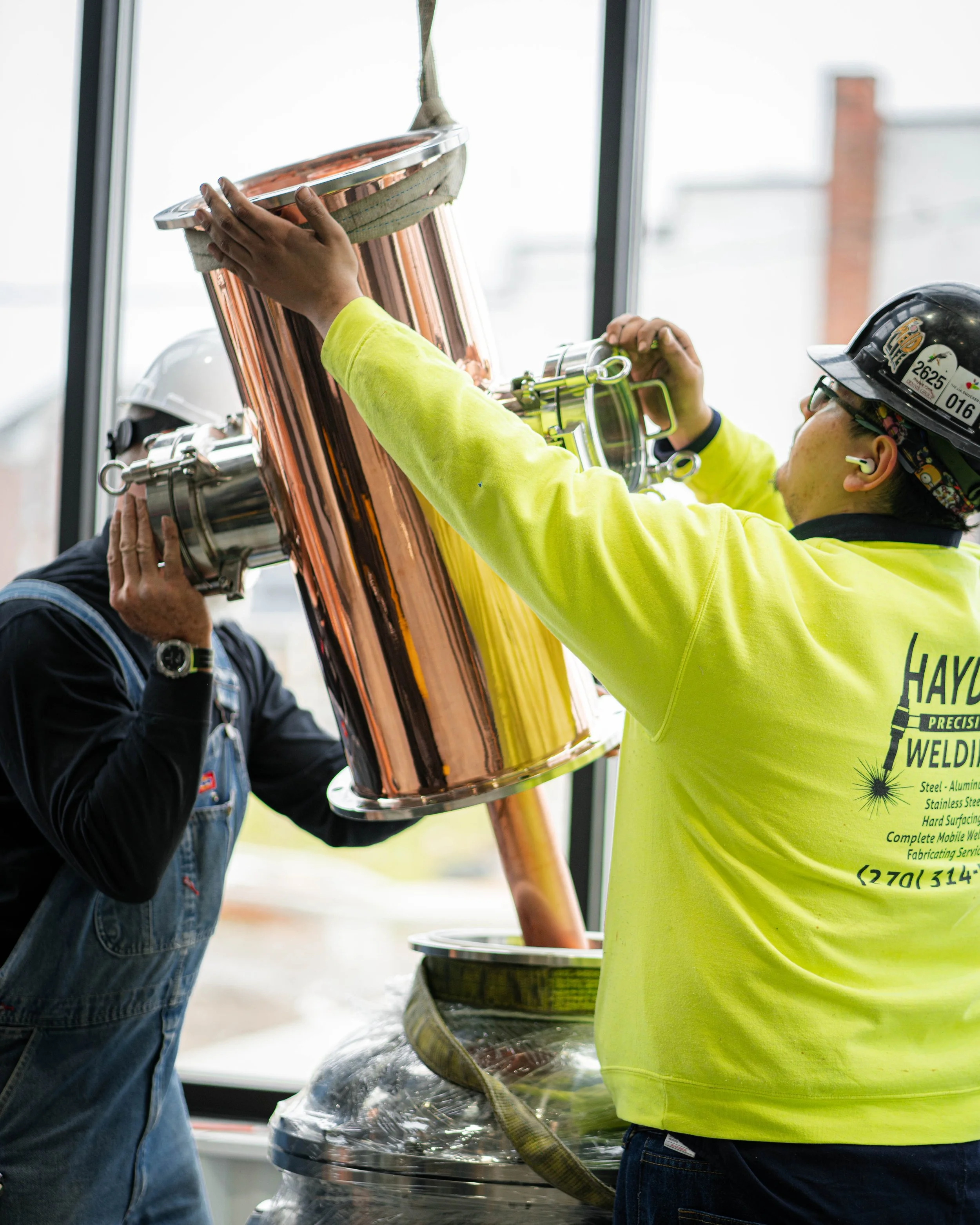 Two workers in safety gear installing a large metal chimney pipe.