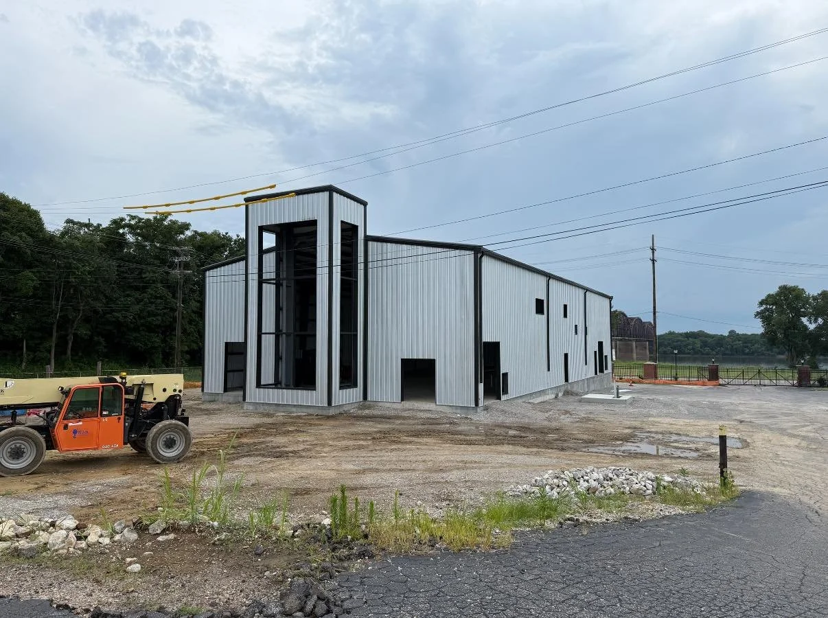 Under construction modern steel building with a tower-like front and large windows, on a dirt lot with construction equipment and power lines in the background.