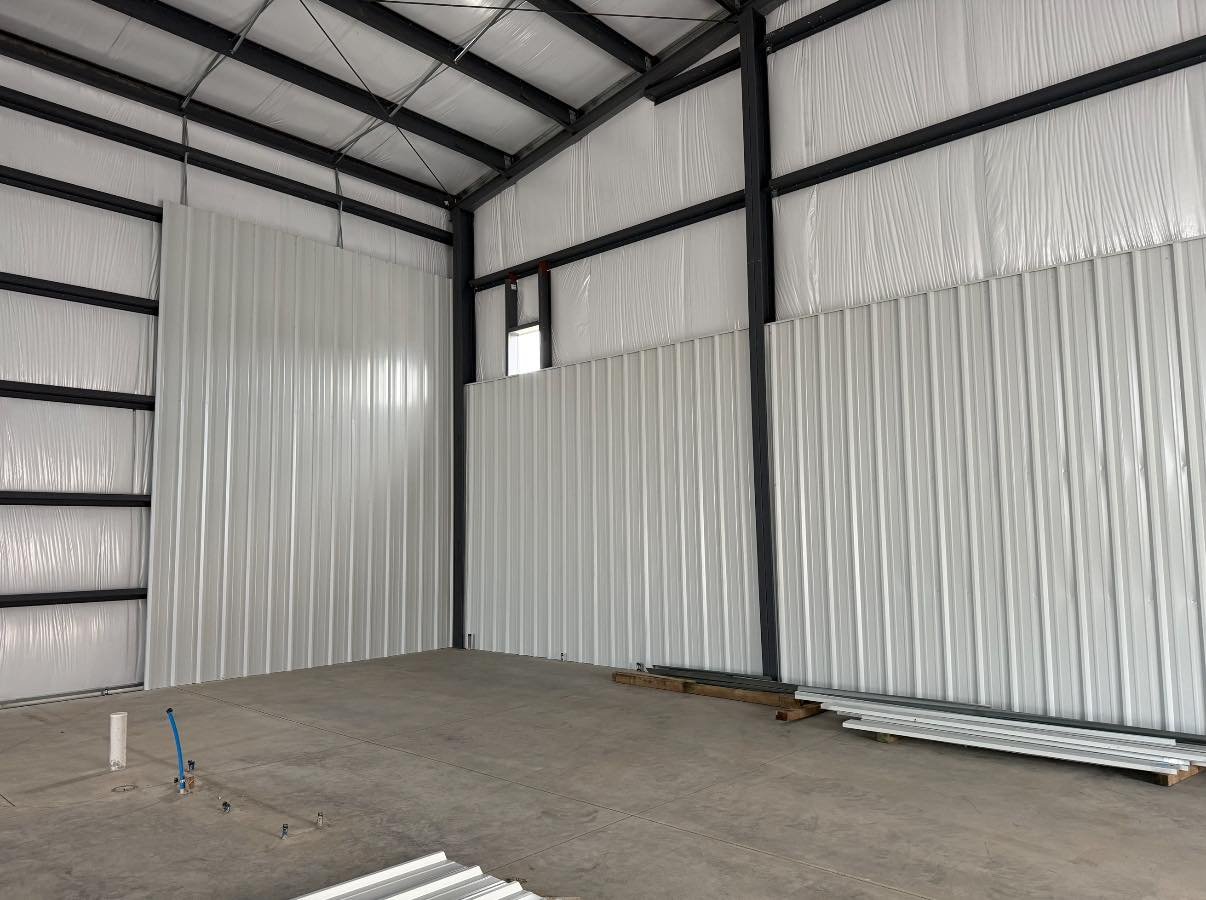 Interior of an empty metal building with unfinished concrete floor, some metal rails and pipes on the floor, and walls made of white corrugated metal panels. The ceiling has a metal framework with some wires.