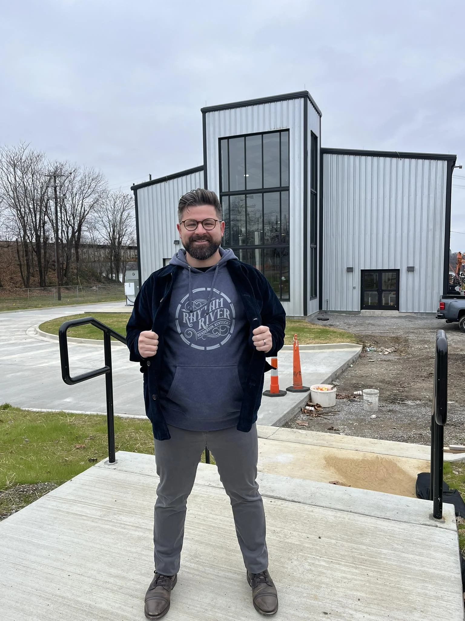 A man with glasses and a beard stands on a concrete sidewalk outside a modern metal building, smiling and holding the collar of his dark jacket. The building has large glass windows and is under construction or renovation, with construction cones and