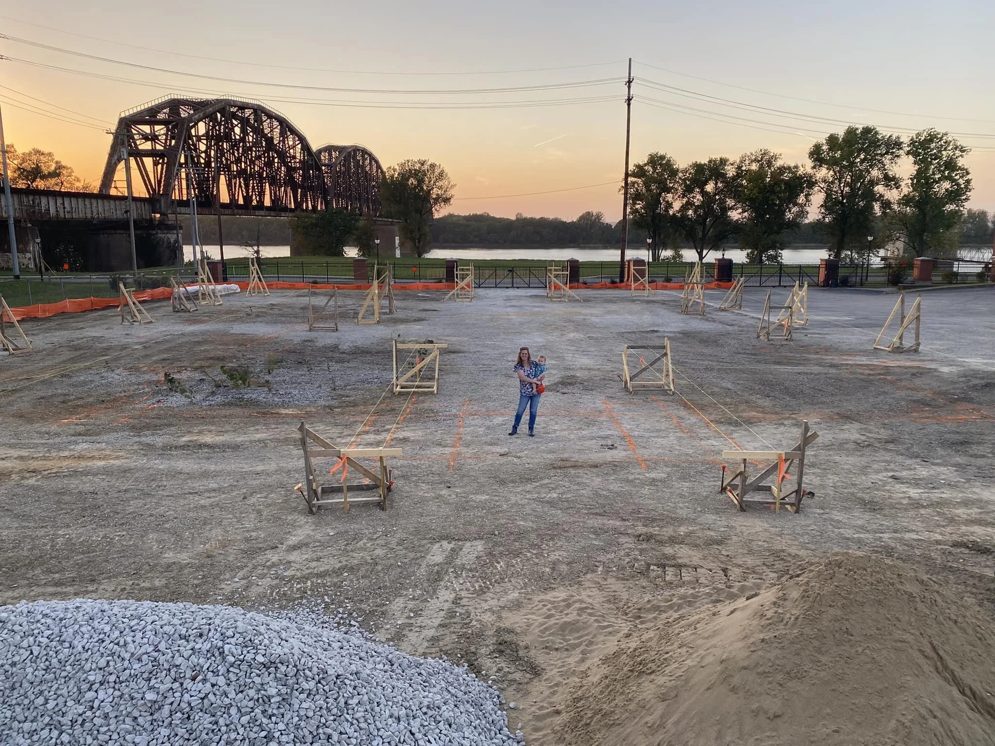 A woman holding a child standing on a construction site in front of a river, with trees and a bridge in the background during sunset.
