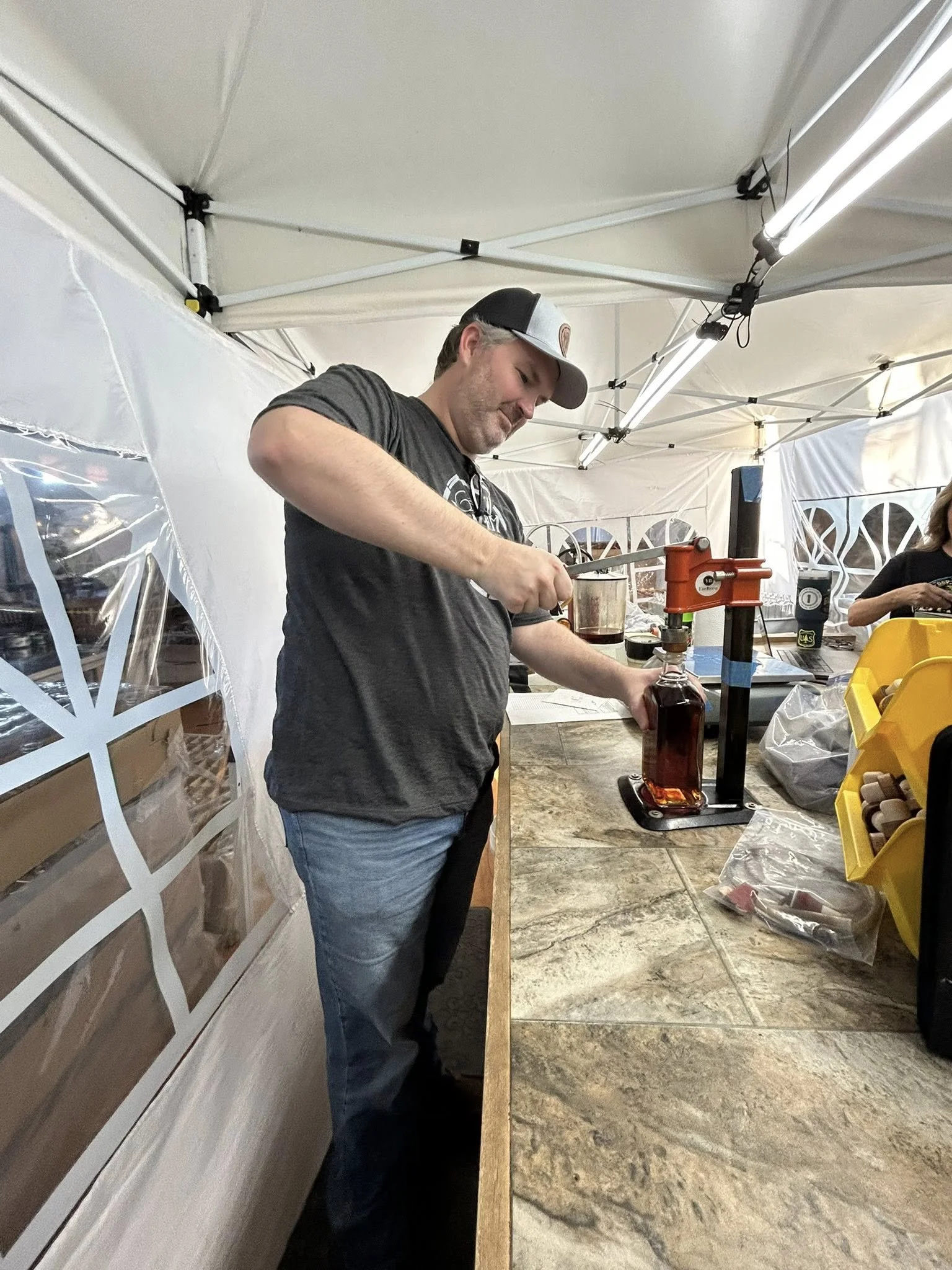 A man wearing a cap and a dark T-shirt is pouring liquid from a measuring cup into a bottle using a bottle capping machine at an outdoor event booth with a white canopy.