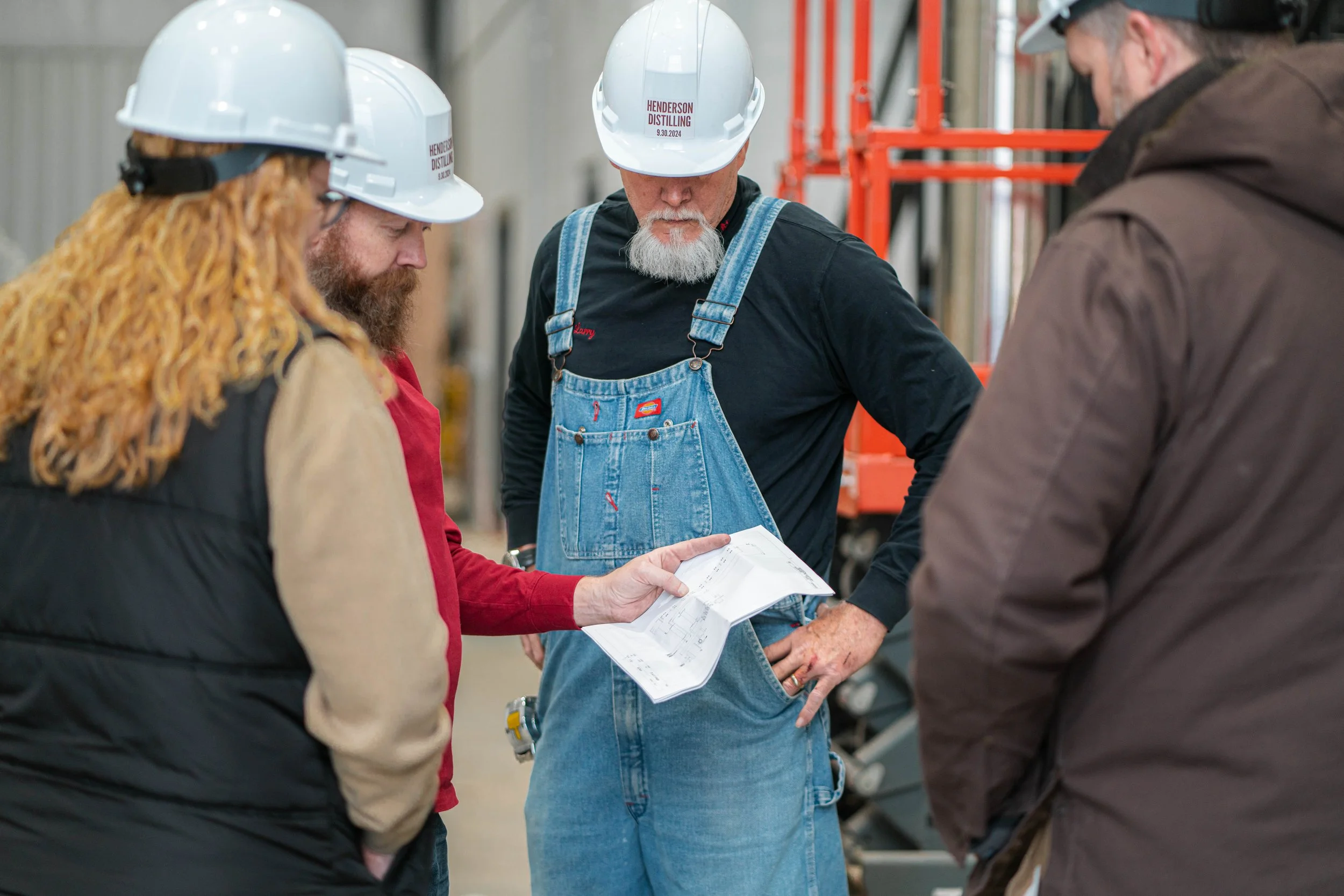 Four workers wearing hard hats and casual work clothes examining blueprints in an industrial setting.