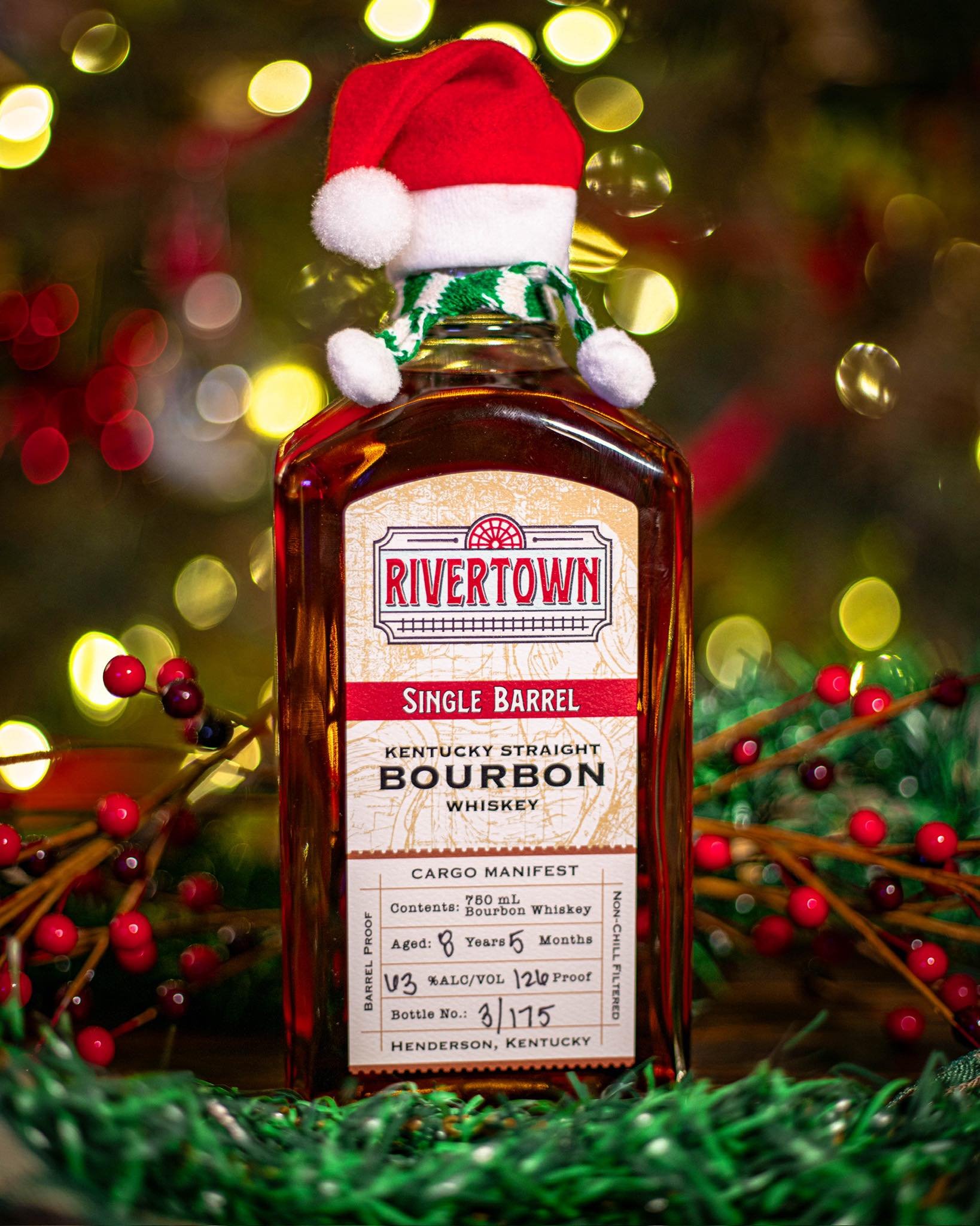 Bottle of Rivertown single barrel bourbon whiskey wearing a festive Santa hat with a green and white scarf, surrounded by Christmas decorations including red berries, green tinsel, and blurred holiday lights in the background.