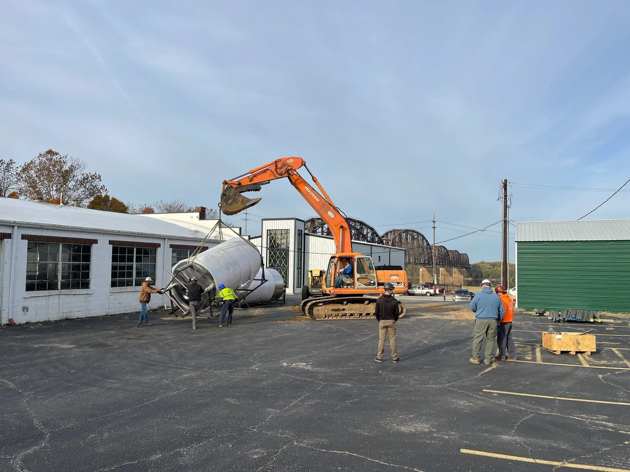 Construction workers guiding a large metal tank into a building with an orange excavator in a parking lot.