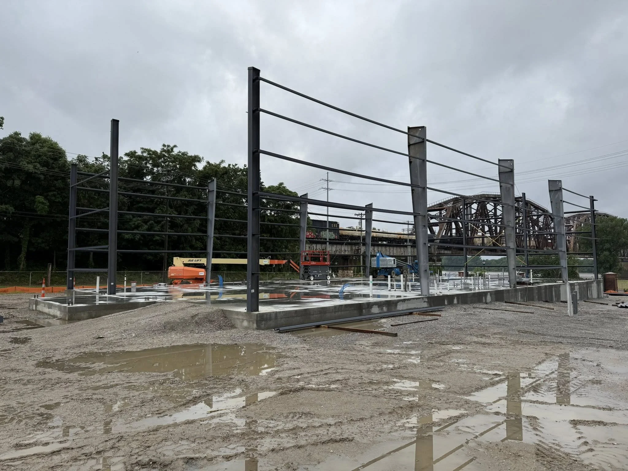 Construction site with steel framework for a building, muddy ground with puddles, construction equipment, and an overcast sky.