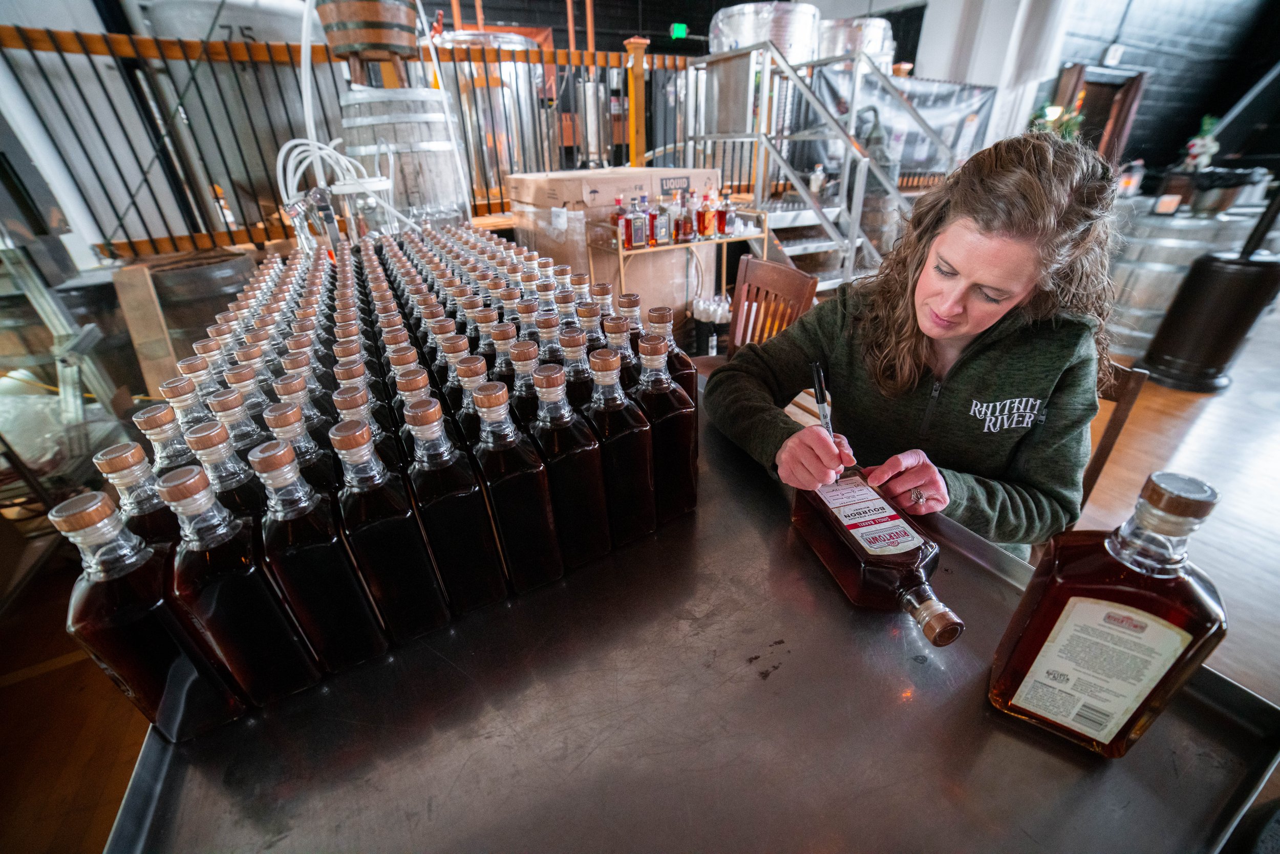 Woman signing a bottle of bourbon at a distillery, with many similar bottles lined up on the table.