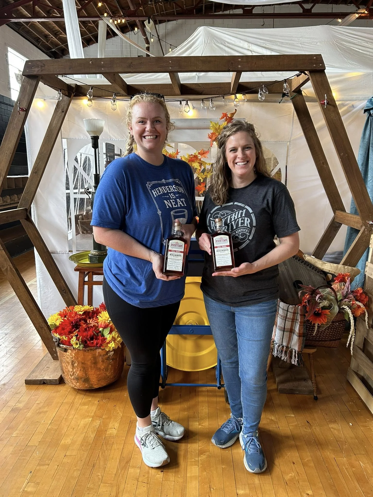 Two women standing inside a booth decorated with autumn-themed flowers and leaves, holding bottles of whiskey and smiling at the camera.