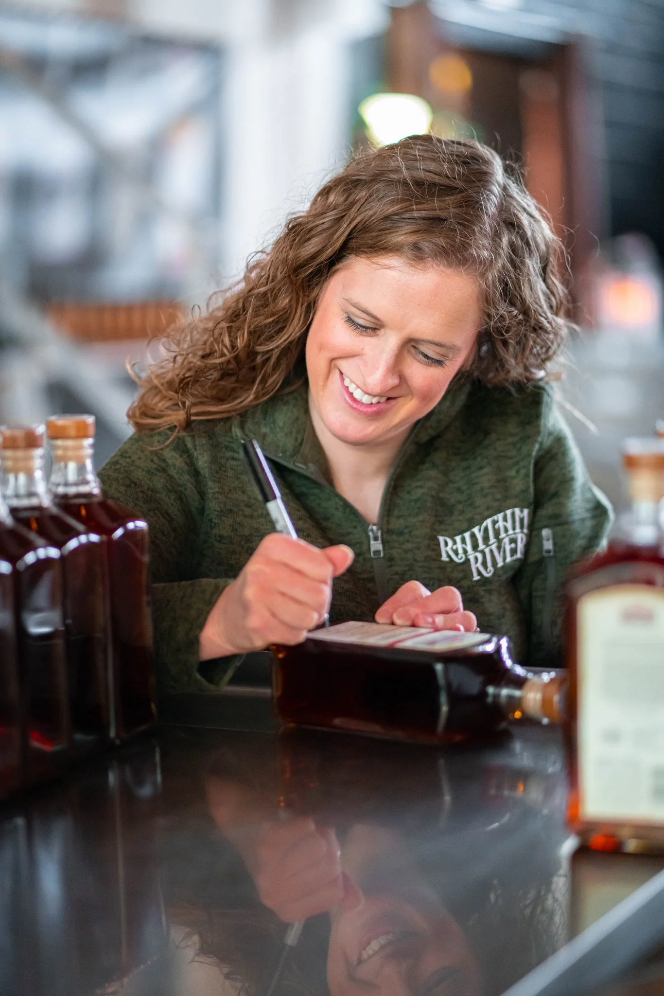 A woman with curly brown hair smiling and writing on a bottle with a black marker at a bar or brewery, wearing a green hoodie with 'Rhythm River' embroidered on it.