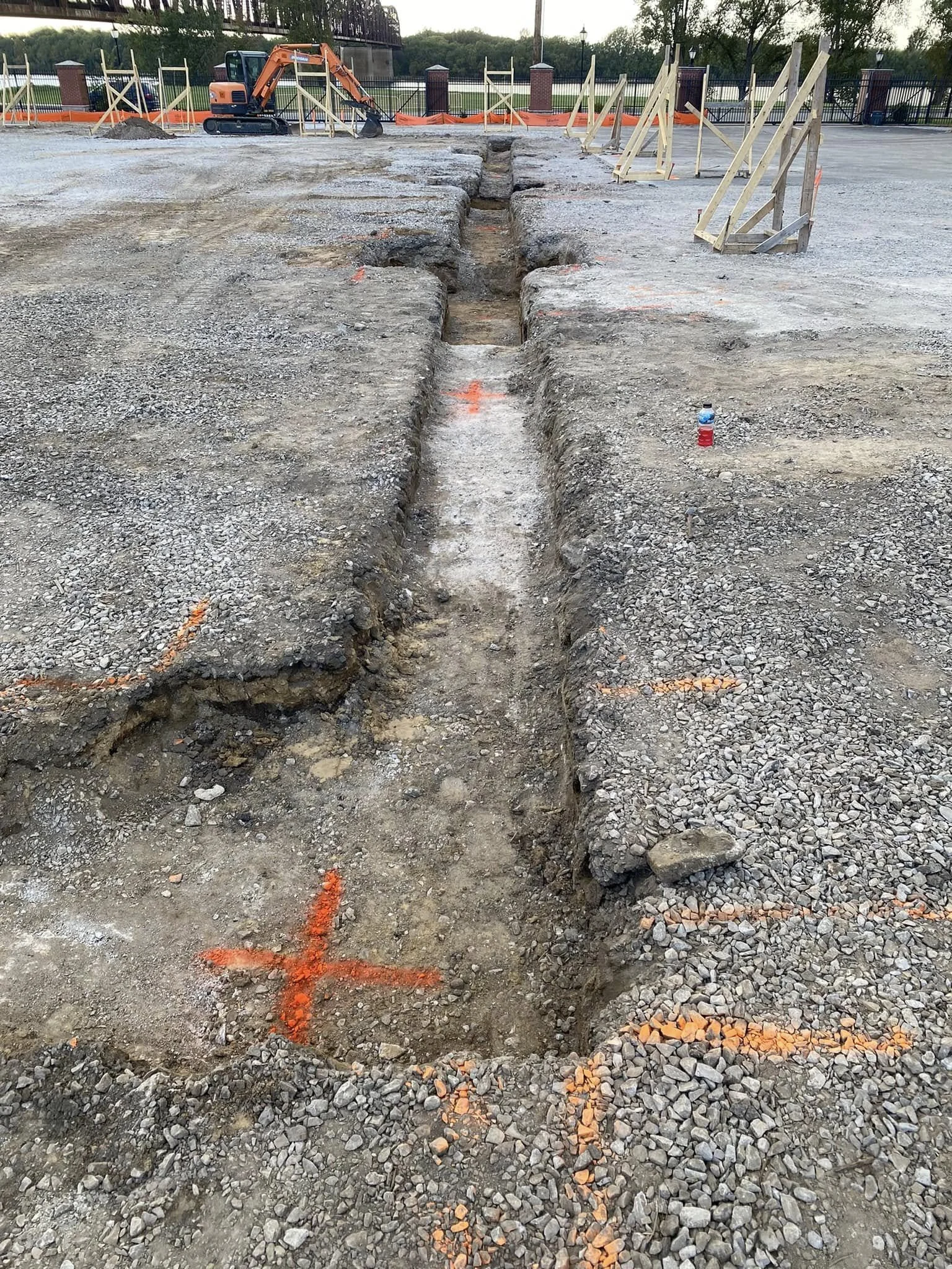 Construction site with a trench for underground utilities, orange markings, a miniature excavator in the background, and wooden barriers on the sides.