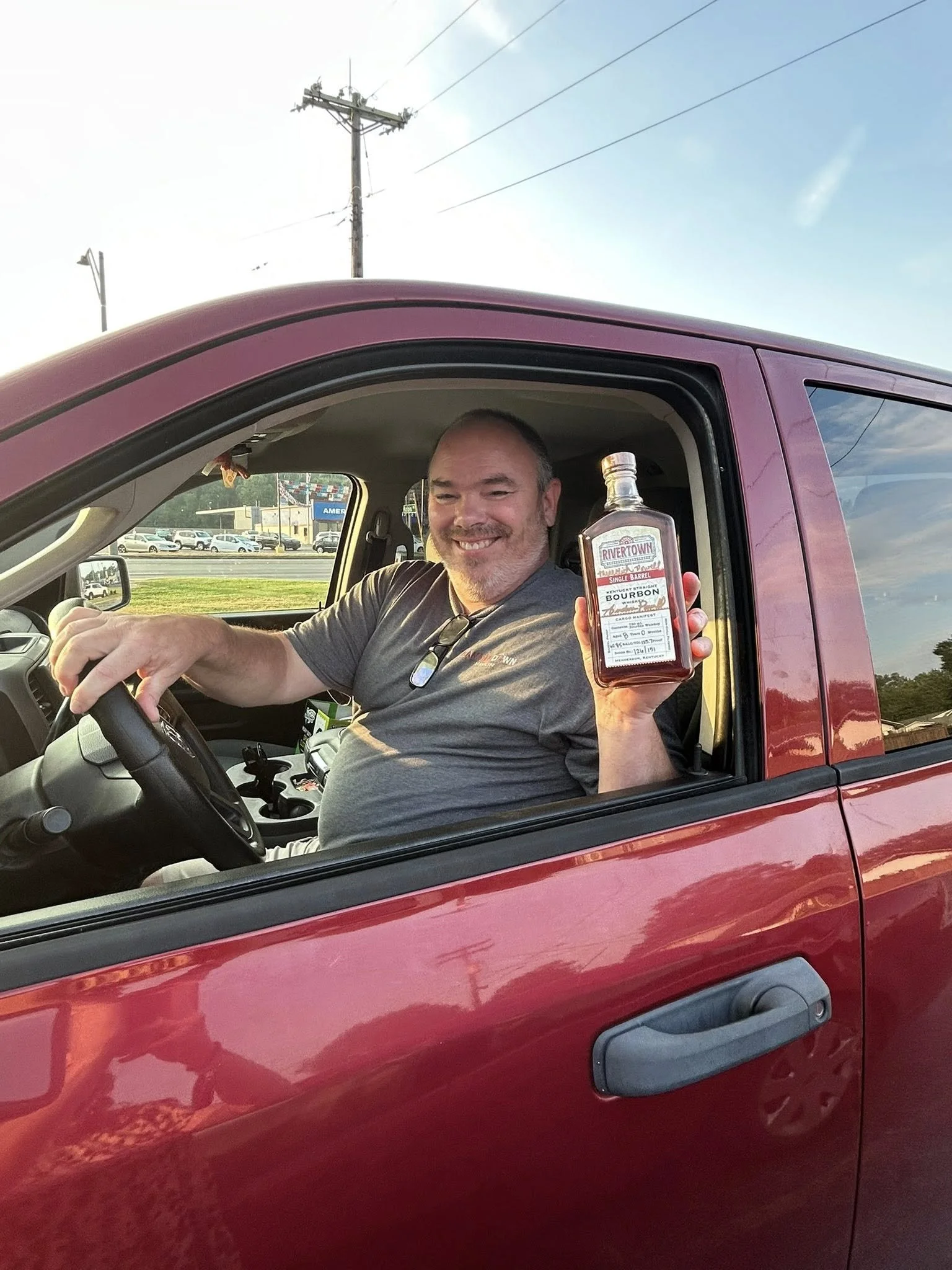 Smiling man sitting in a red pickup truck, holding a bottle of bourbon, with a parking lot and power lines visible outside.