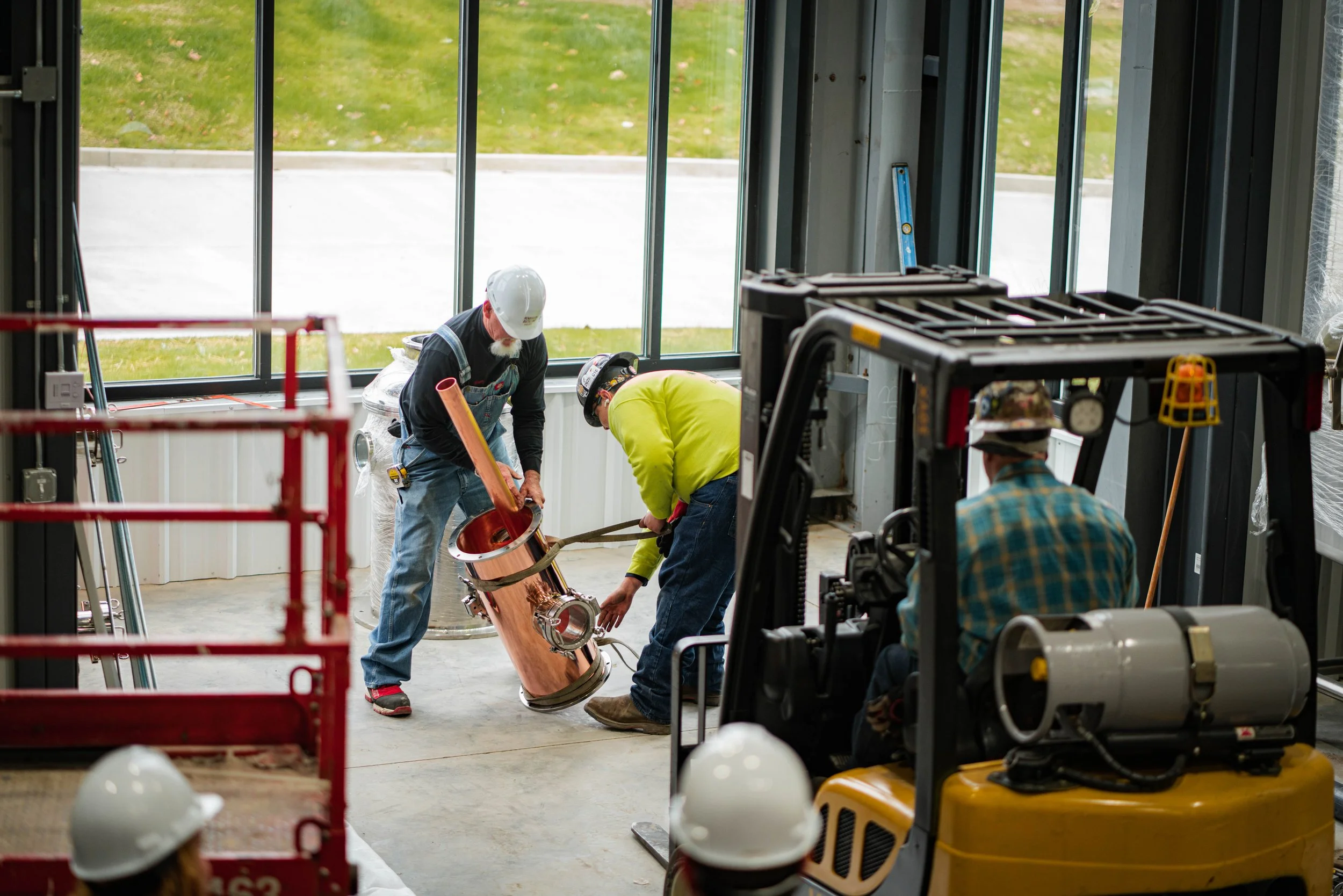 Three construction workers in safety gear working on a copper pipe inside a building, with large windows and construction equipment nearby.