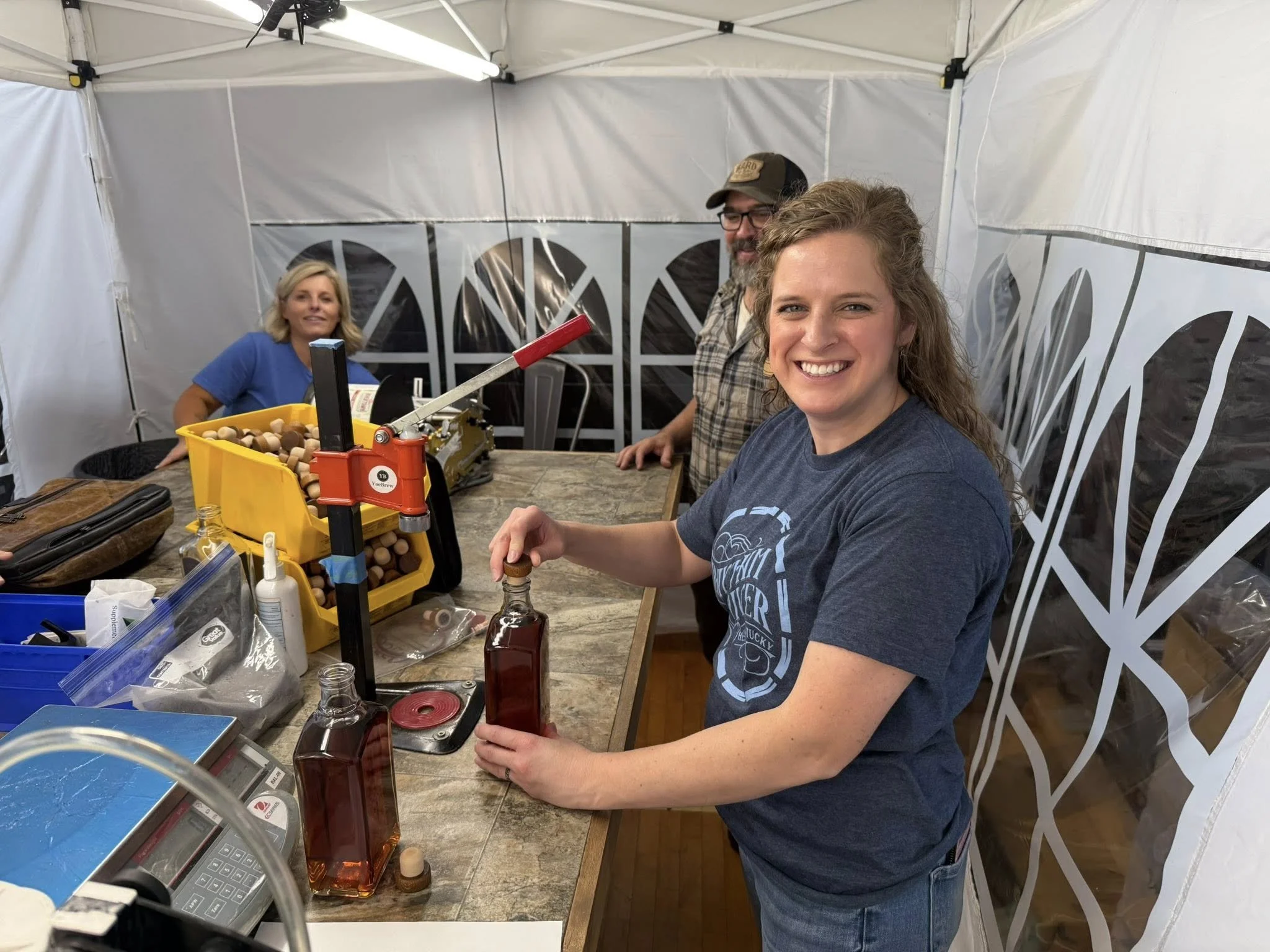 Three people working at a wooden table inside a white tent, with merchandise, bottles, and equipment, smiling and posing for the photo.