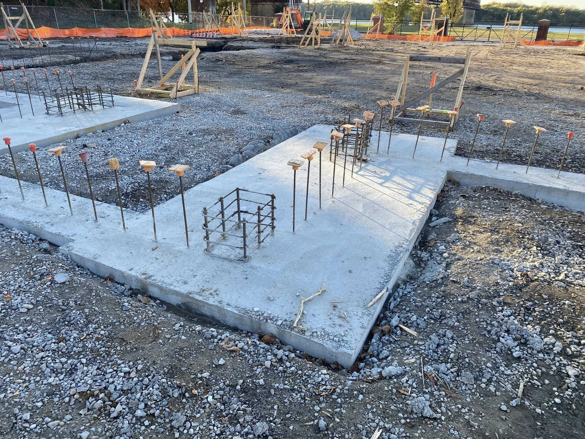 Construction site with concrete foundation and metal rebar protruding from it, surrounded by gravel, with wooden forms and orange safety fencing in the background.