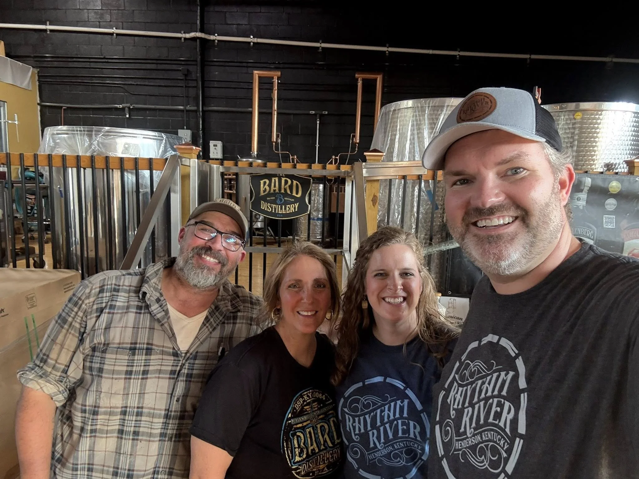 Four smiling people taking a selfie inside a distillery with copper stills in the background. The group includes two men and two women, all wearing casual clothing and branded shirts.