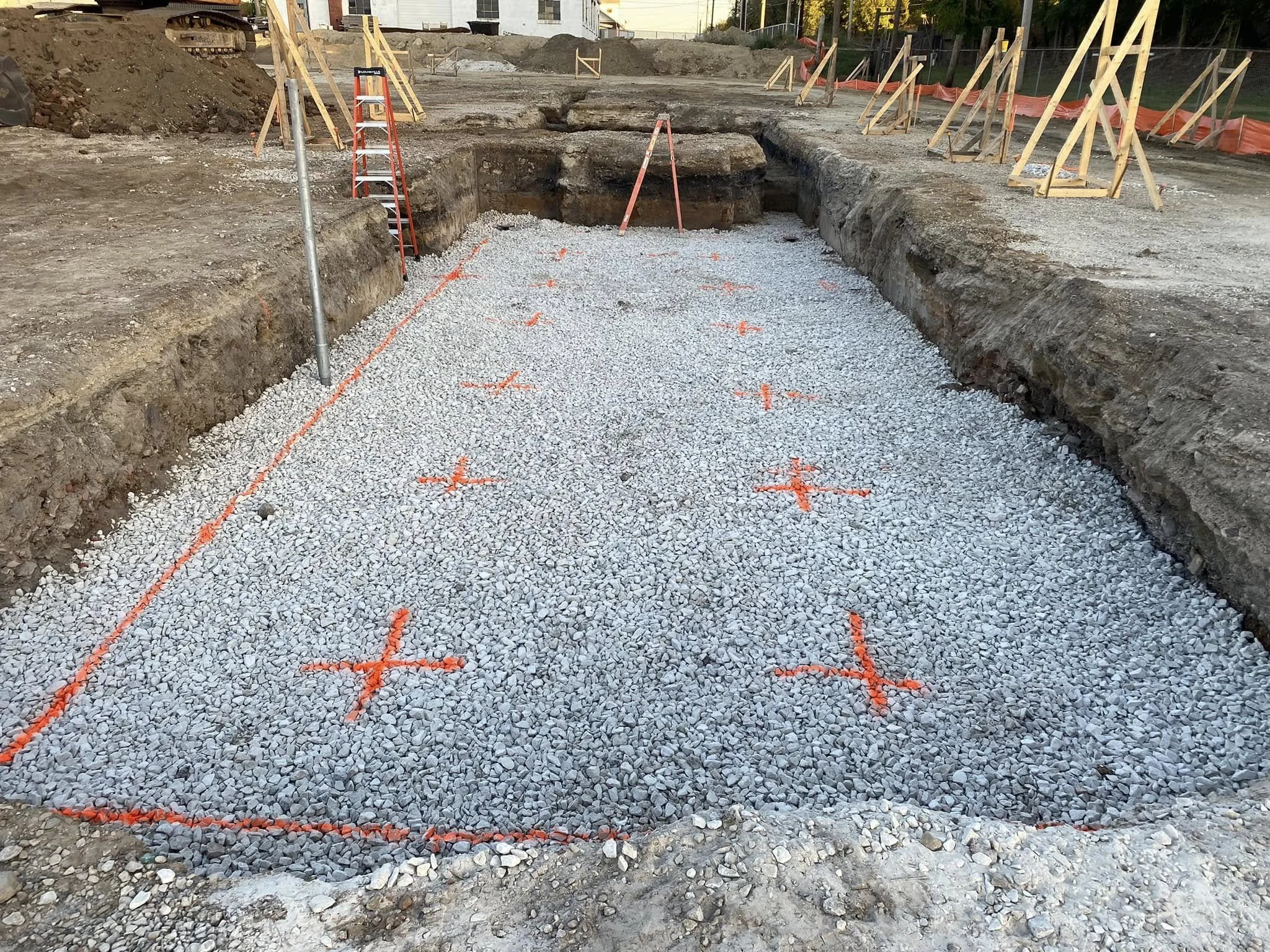 Construction site with excavated area filled with gravel, marked with orange spray paint for foundation layout, wooden formwork on edges, and construction equipment and materials in background.