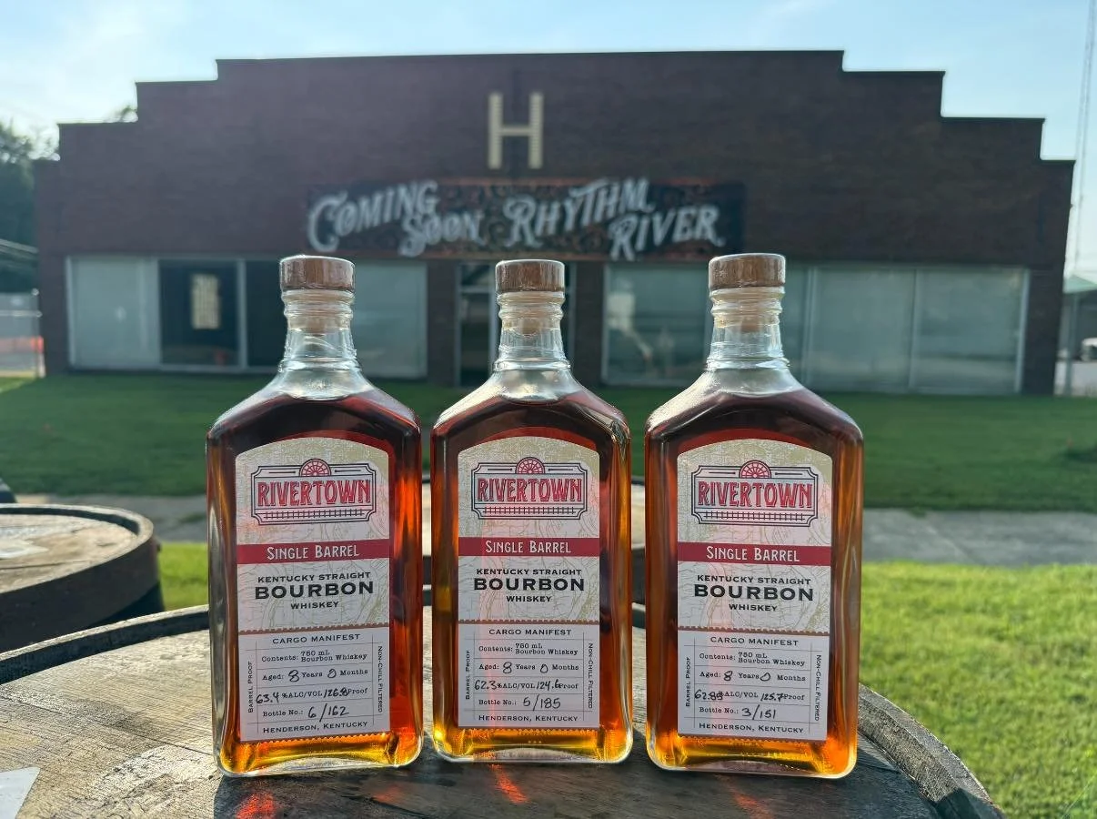 Three bottles of Rivertown single barrel Kentucky straight bourbon whiskey placed on a wooden surface outdoors, with a brick building with a sign that reads "Coming Soon Rhythmin River" in the background.