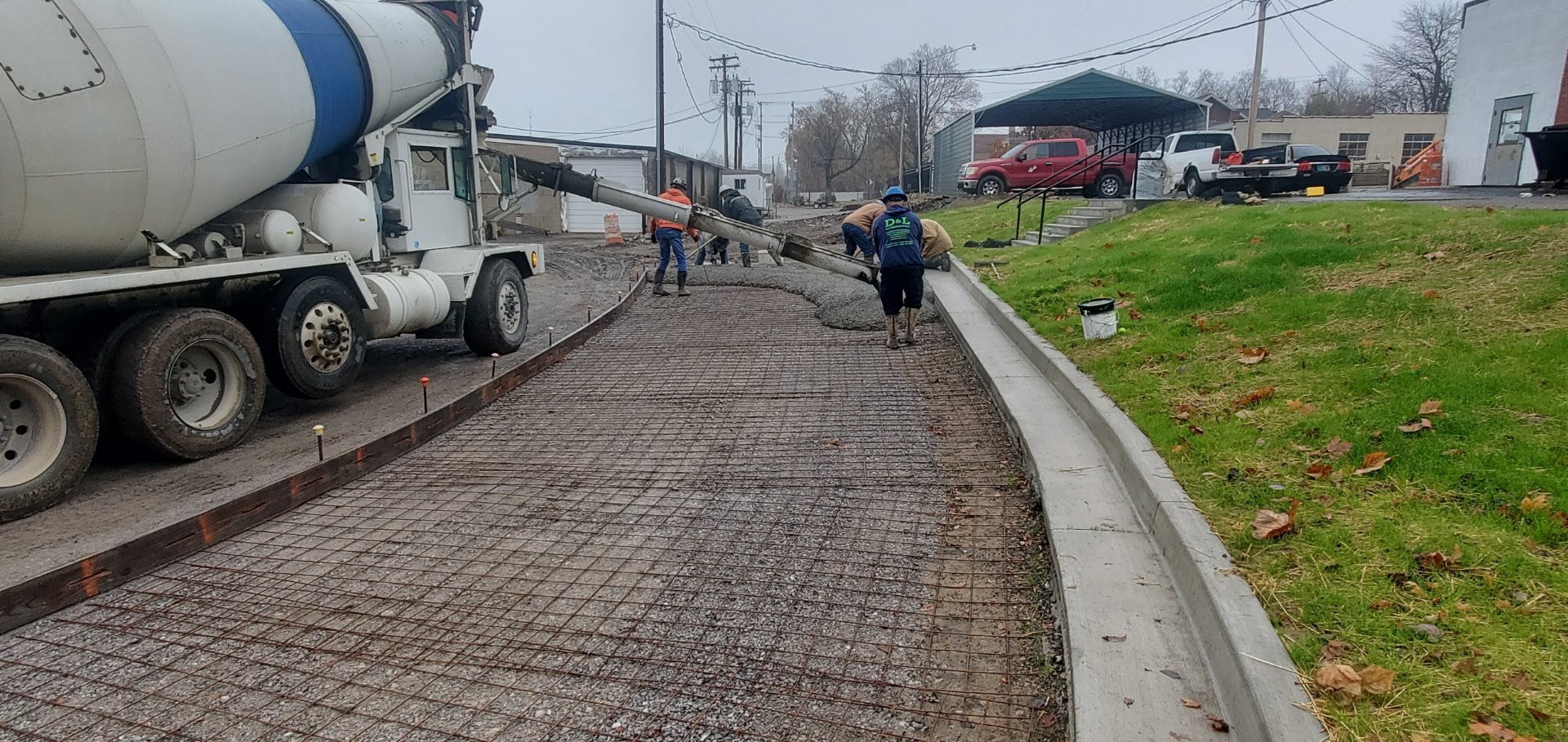 Workers are pouring and smoothing concrete on a curved sidewalk with a cement mixer truck nearby, over a wire mesh reinforcement. There is grass and a small staircase with a handrail on the right side of the sidewalk. Several parked cars and building