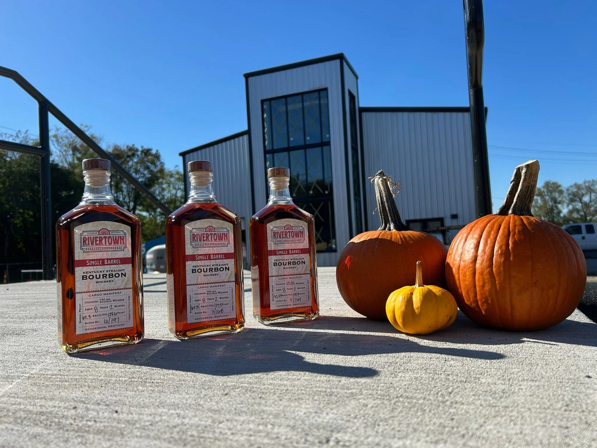 Three bottles of Rivertown Kentucky Straight Bourbon Whiskey and three pumpkins of varying sizes and colors on a table outdoors with a blue sky and modern building in the background.