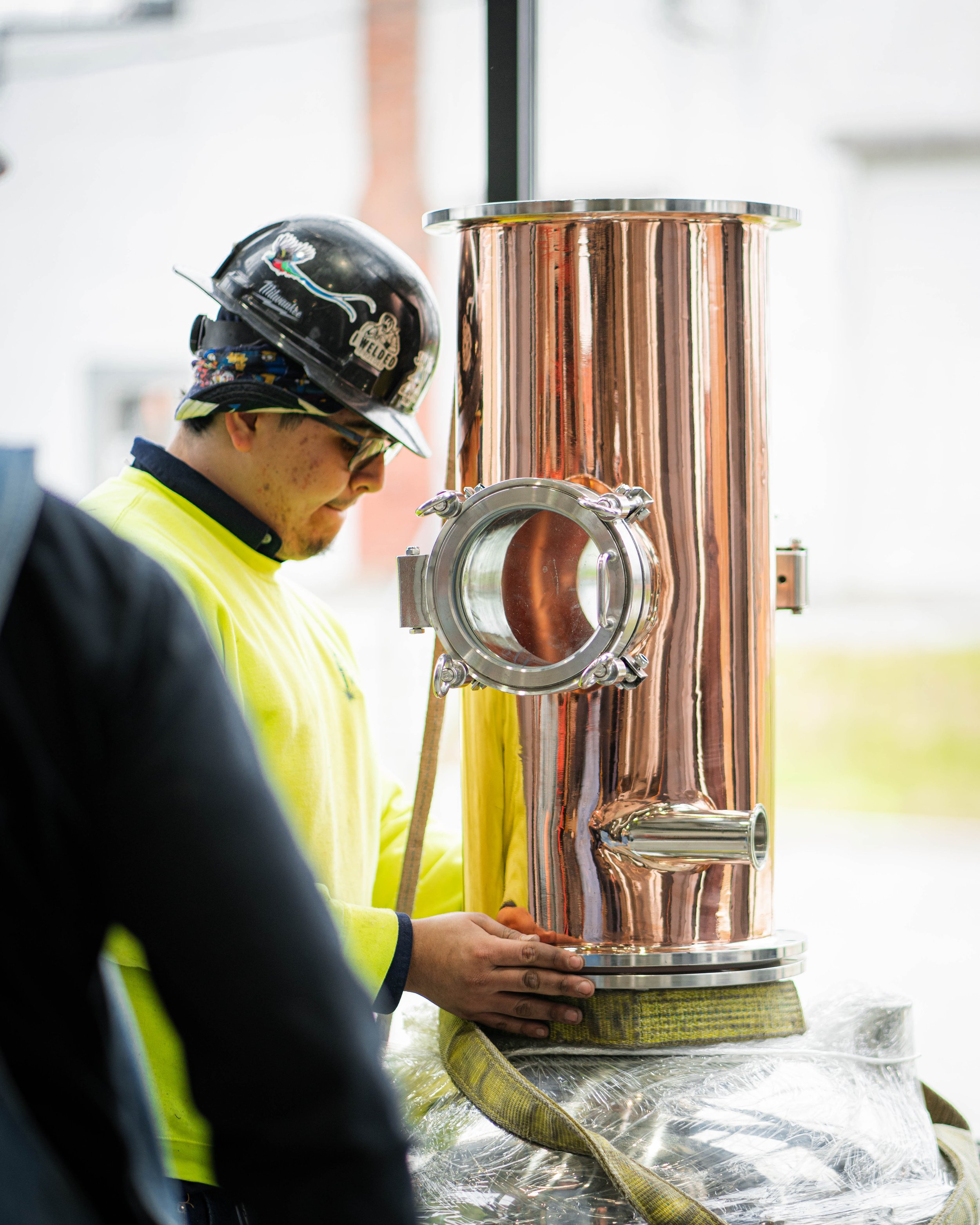 A worker in a yellow safety shirt and a helmet inspects a large copper brewing vessel with a small side window.