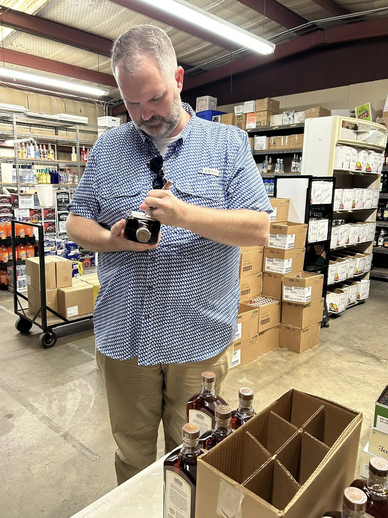 A man with short hair and a beard wearing a blue patterned button-up shirt and khaki pants is in a warehouse or store, holding a small bottle and inspecting it closely. There are several bottles of alcohol on the table in front of him and shelves wit