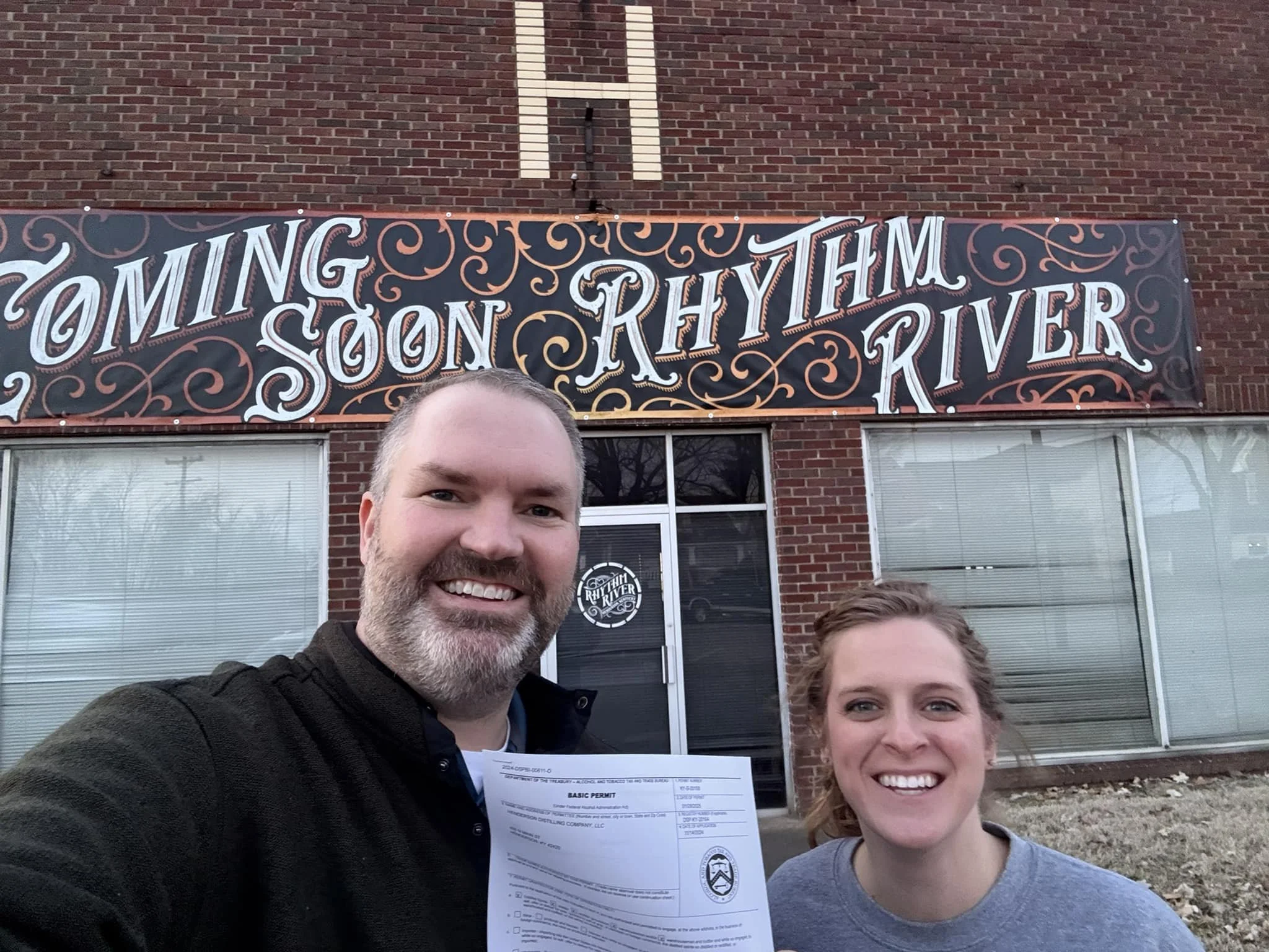 Two smiling people taking a selfie in front of a brick building with a sign that reads 'Coming Soon Rhythm River.' One person is holding a permit.