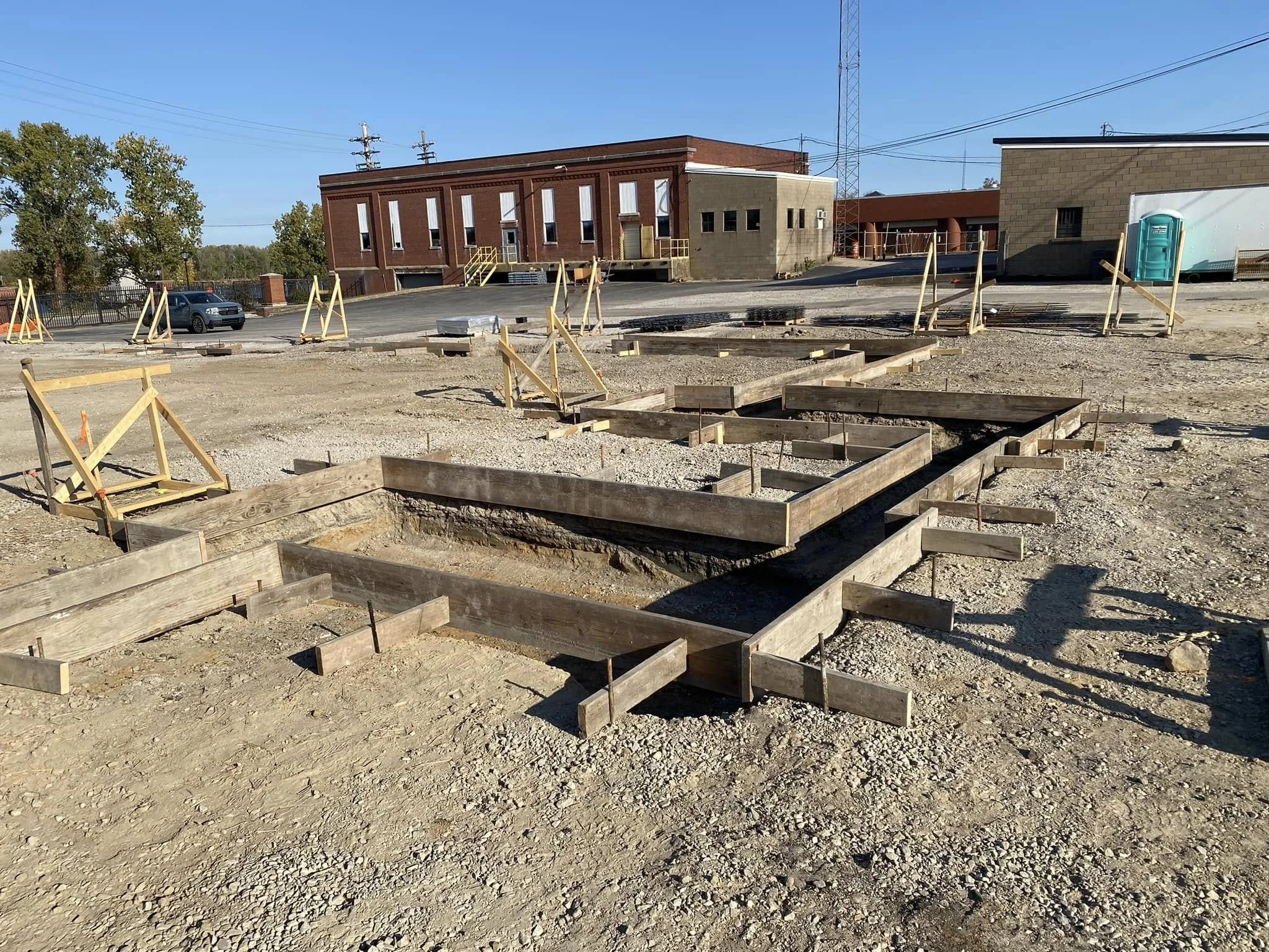 Construction site with wooden framing for a building foundation, surrounded by gravel, with a brick building, a portable toilet, and power lines in the background.