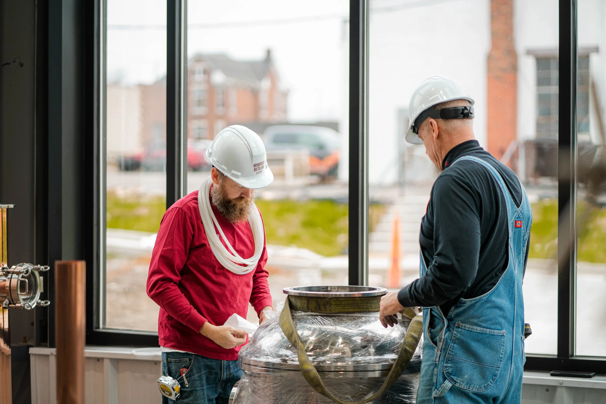 Two construction workers wearing hard hats inside a building, working with a large metal container covered with plastic.