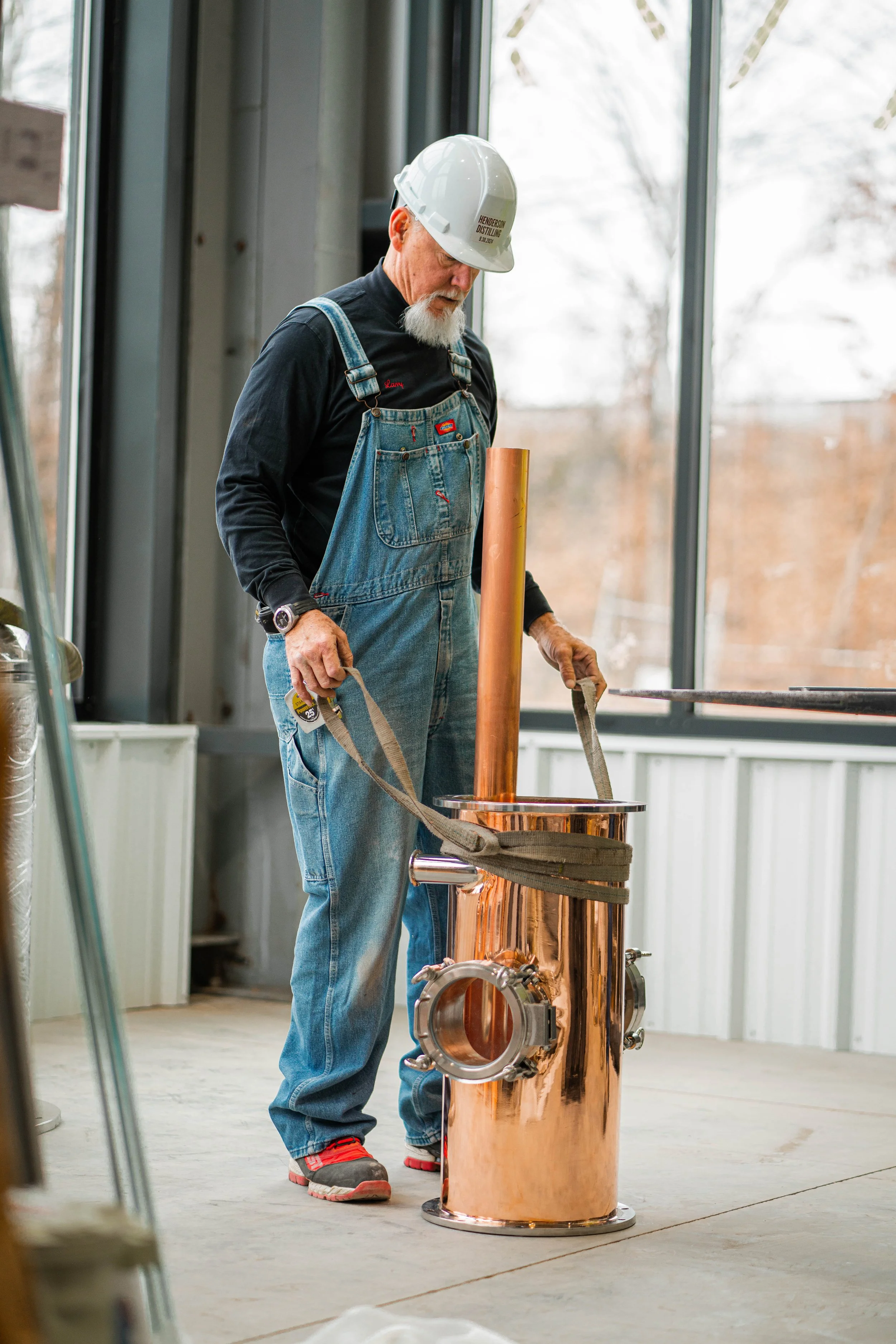 A man wearing a white hard hat, black shirt, and blue overalls stands next to a copper distillation apparatus placed inside a room with large windows. The man appears to be securing or adjusting the equipment.