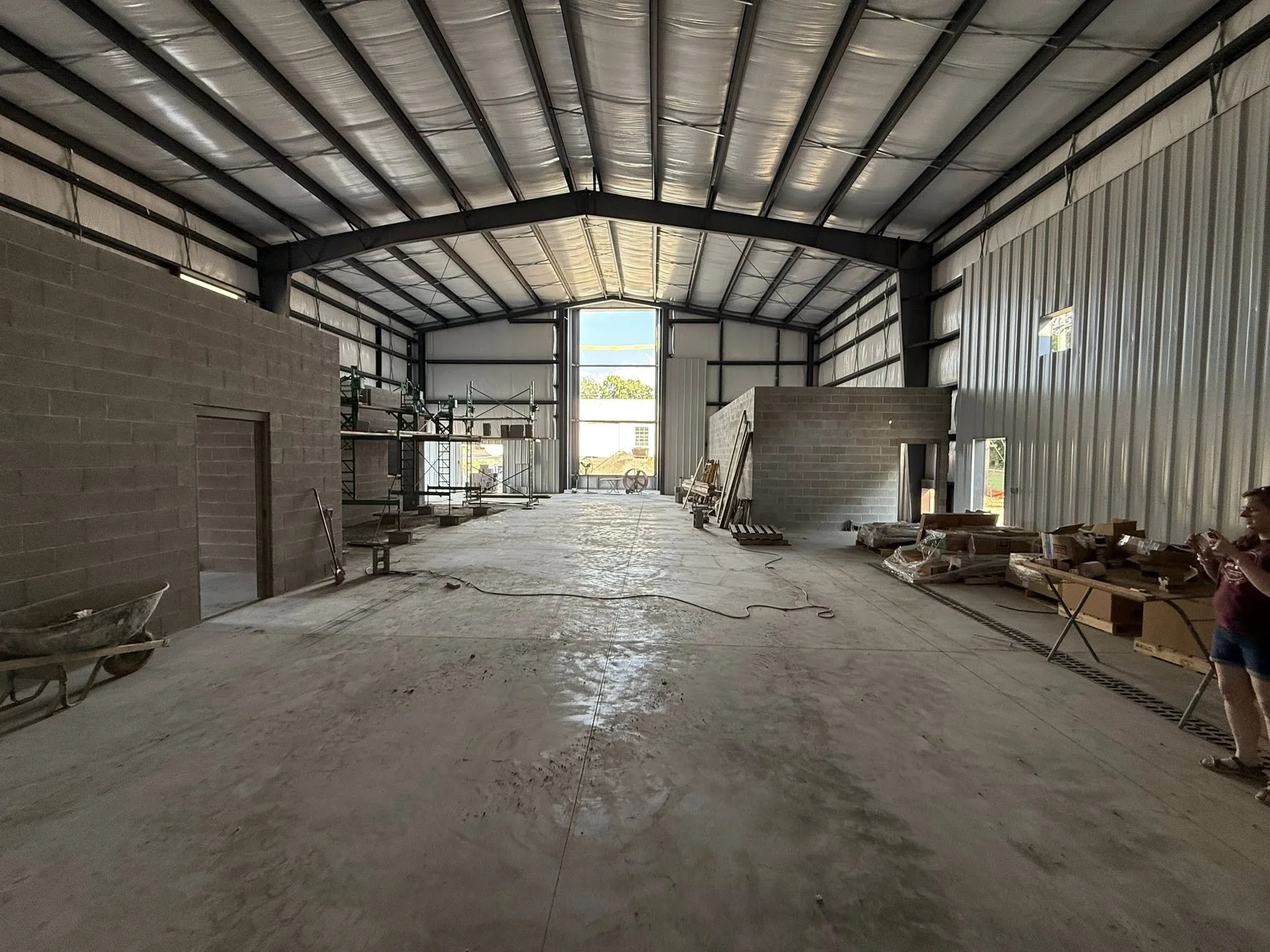 Interior of a warehouse or industrial building under construction, with scaffolding, construction materials, and a person standing on the right taking a photo.