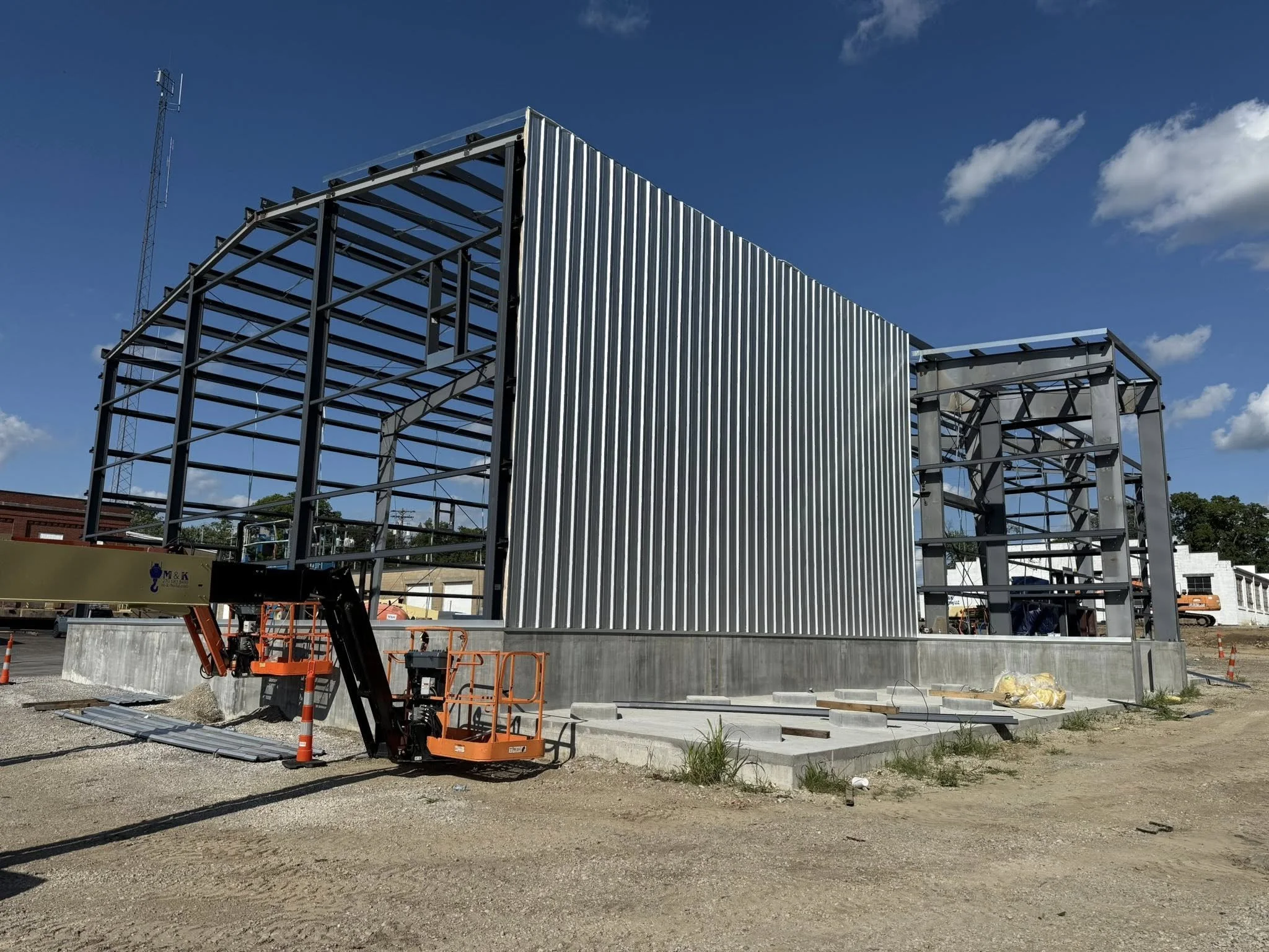 Construction site with steel framework and metal siding in progress, orange lift equipment, gravel ground, partly cloudy sky.