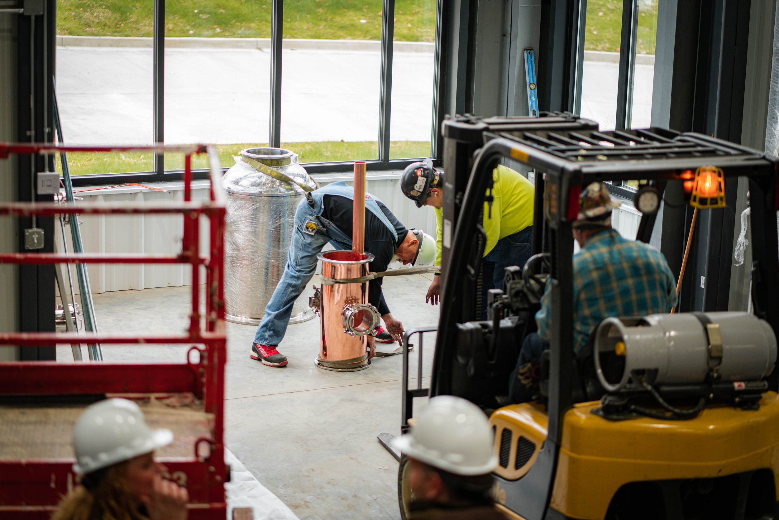 Construction workers installing a copper pipe in an indoor space with large windows, a forklift nearby, and a large metal water tank in the background.