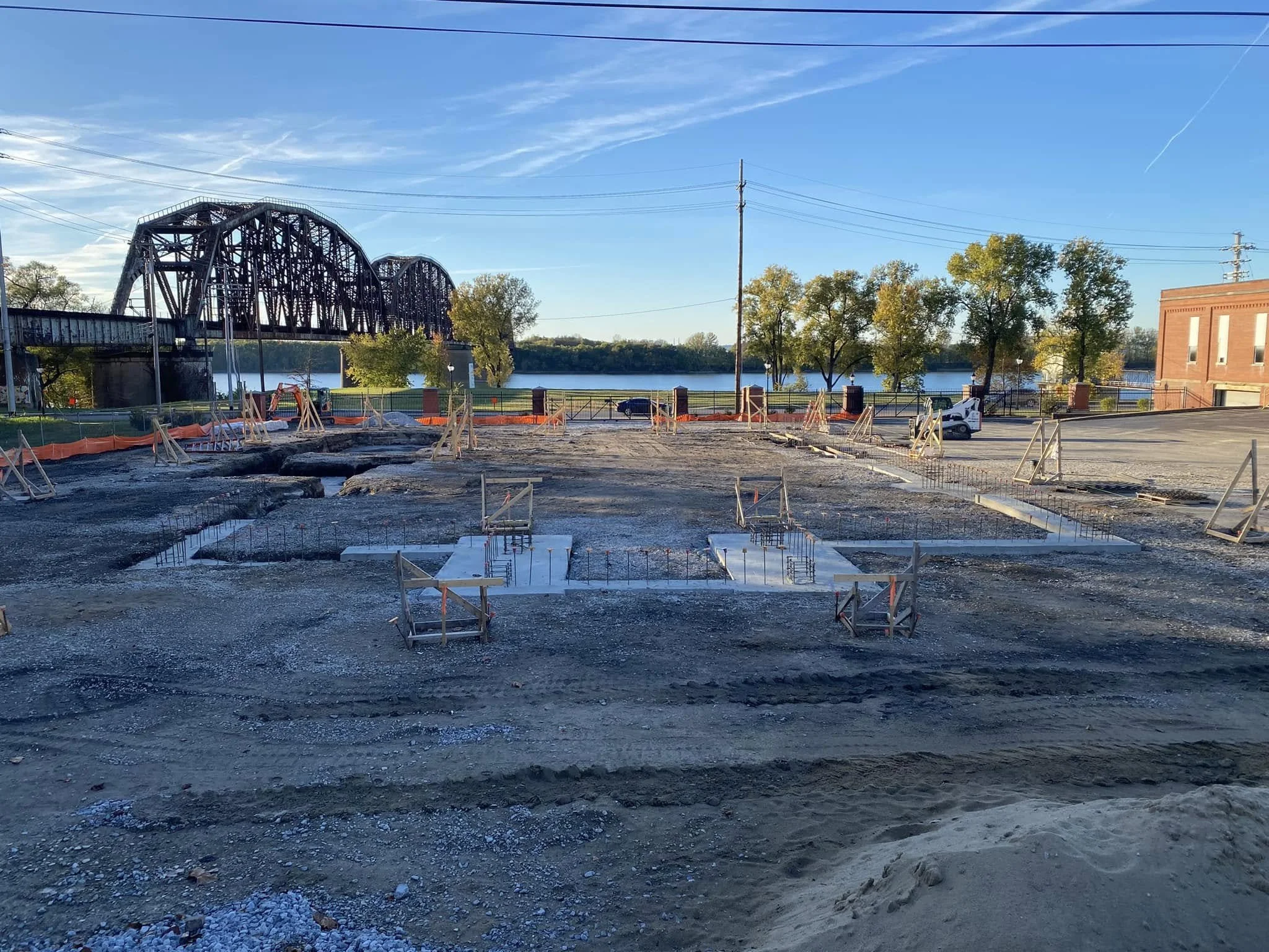 Construction site with concrete pouring forms, with a bridge, river, trees, parked car, a van, and a brick building in the background on a clear day.