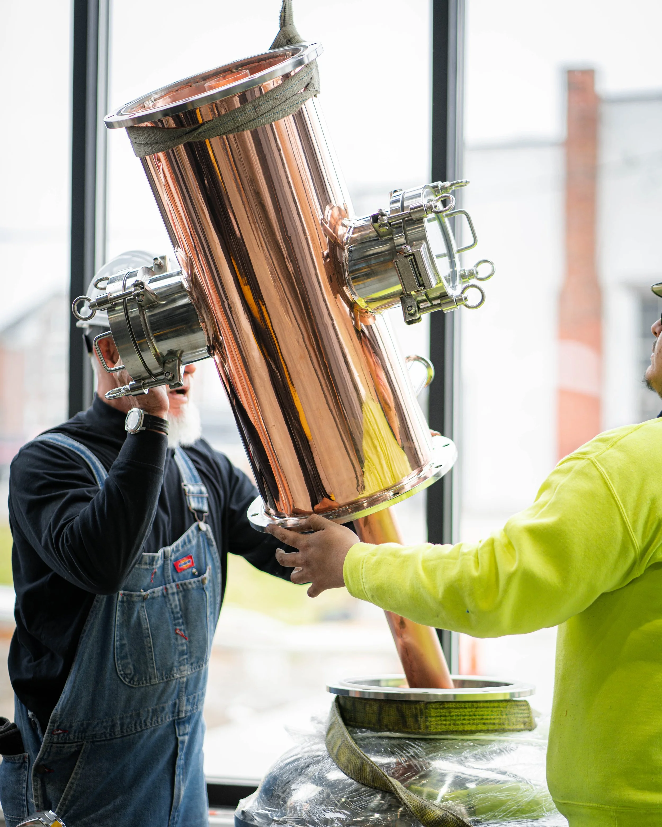 Two men are installing a shiny copper component, possibly part of an industrial or scientific apparatus, in a bright room with large windows.