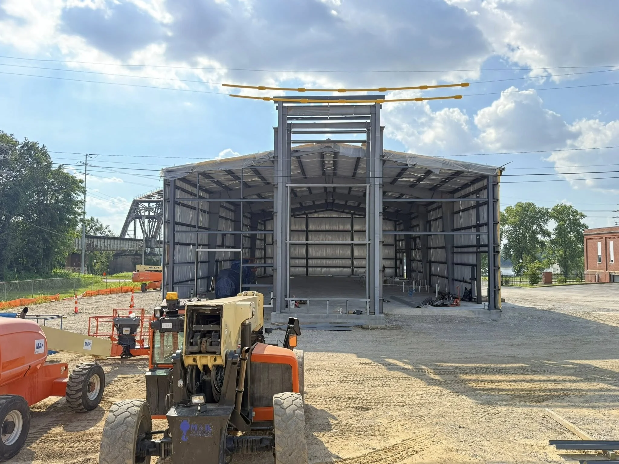 Construction site with a metal frame building in progress, construction equipment, and a partly cloudy sky.